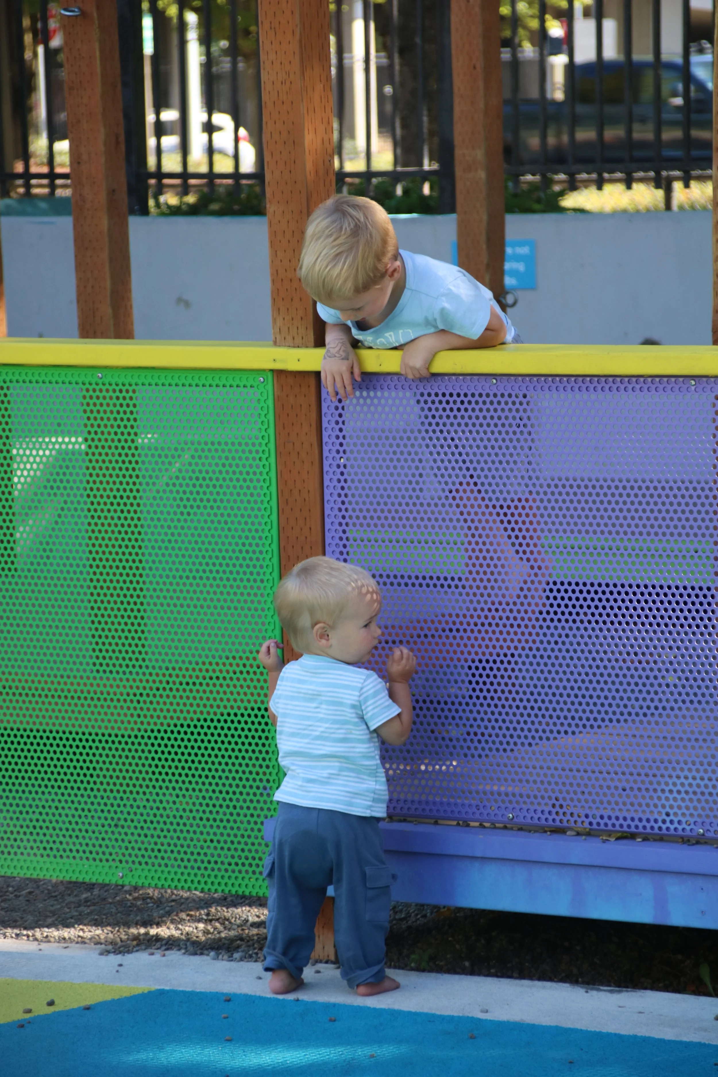 Two young boys, one leaning over a colorful playground barrier and the other standing at its base, engaging with each other at a playground.