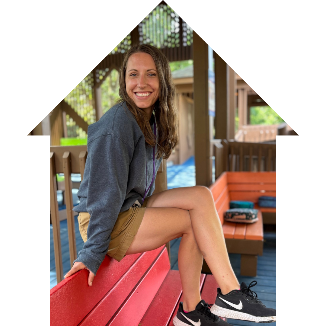 A smiling young woman with long brown hair, wearing a gray sweater, khaki shorts, and black Nike sneakers, sitting on a red wooden bench at an outdoor park or playground.