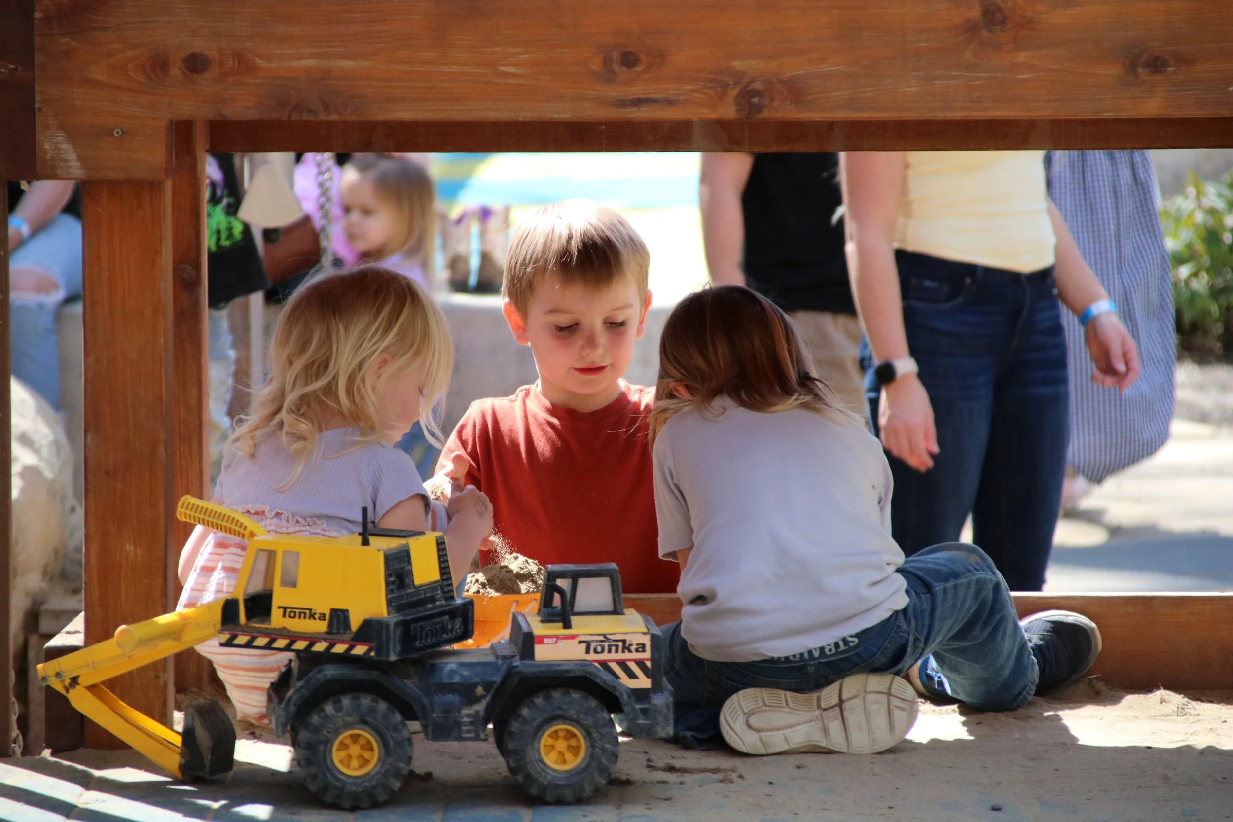 Three young children playing in a sandbox at a park, with a yellow toy excavator nearby, under a wooden structure, while adults and other children are visible in the background.