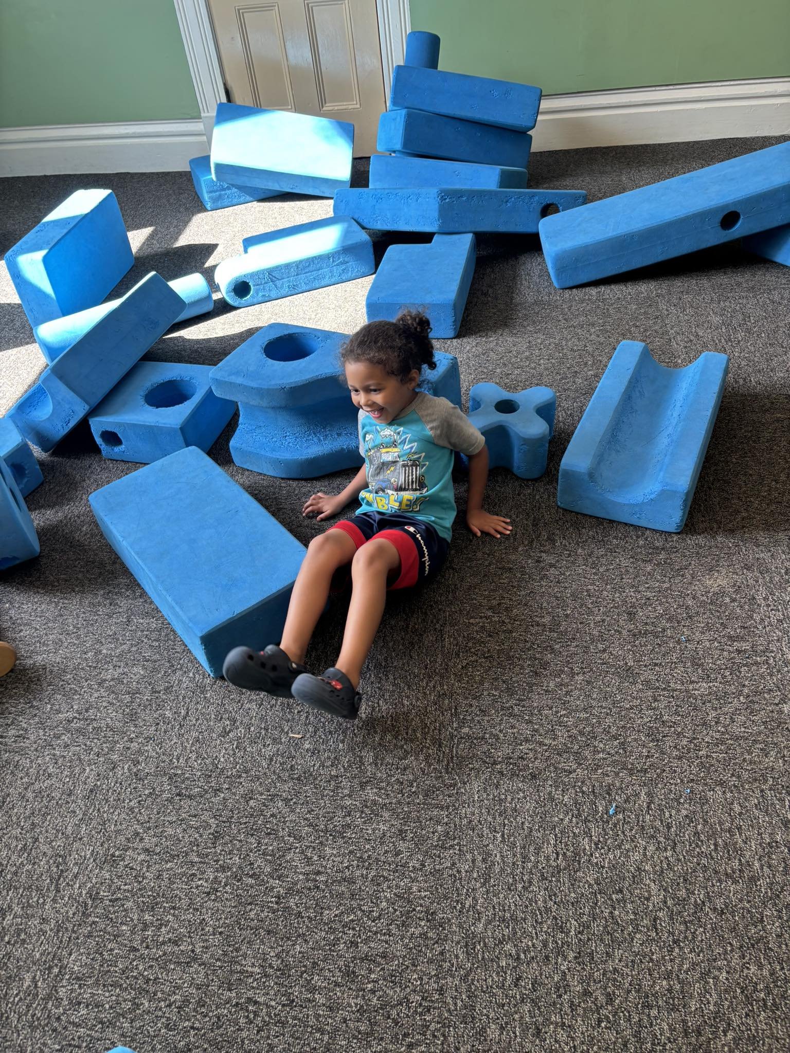 A young boy sitting on the carpeted floor of an indoor playground surrounded by large blue foam blocks and shapes, smiling and enjoying playtime.