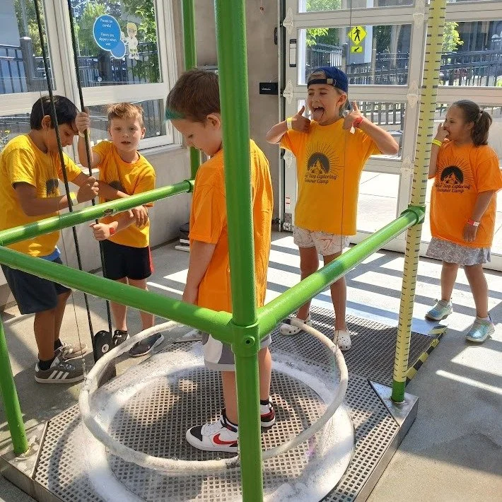 Children playing on a bubble-making station in a sunlit indoor area, wearing orange shirts and casual clothes.