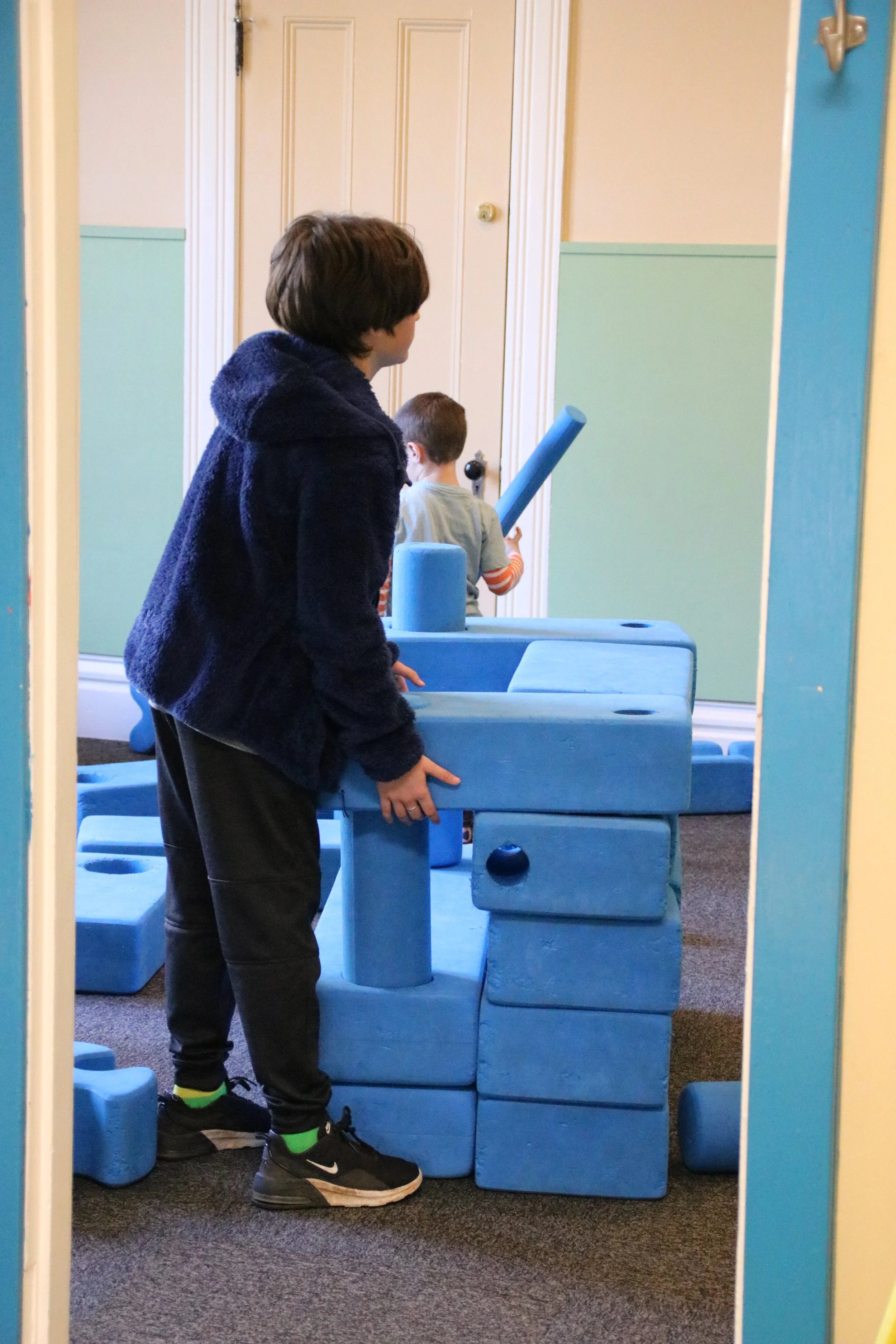Two boys playing with large blue foam blocks in a playroom or indoor area.