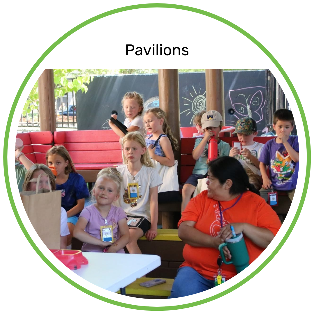 Group of children sitting on a red and yellow bench at a pavilion during daytime, with a woman in an orange shirt in front, some children wearing badges and holding drinks or phones, blackboard with drawings in background.