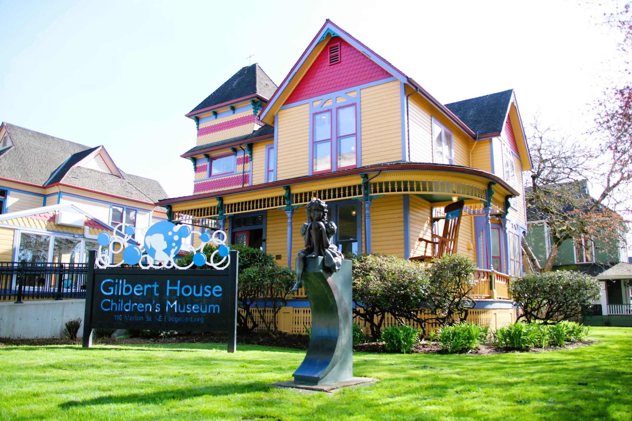 Exterior view of the brightly painted yellow and red trim historic Gilbert House, which serves as the museum entrance, featuring the Gilbert House Children's Museum sign and a bronze sculpture.