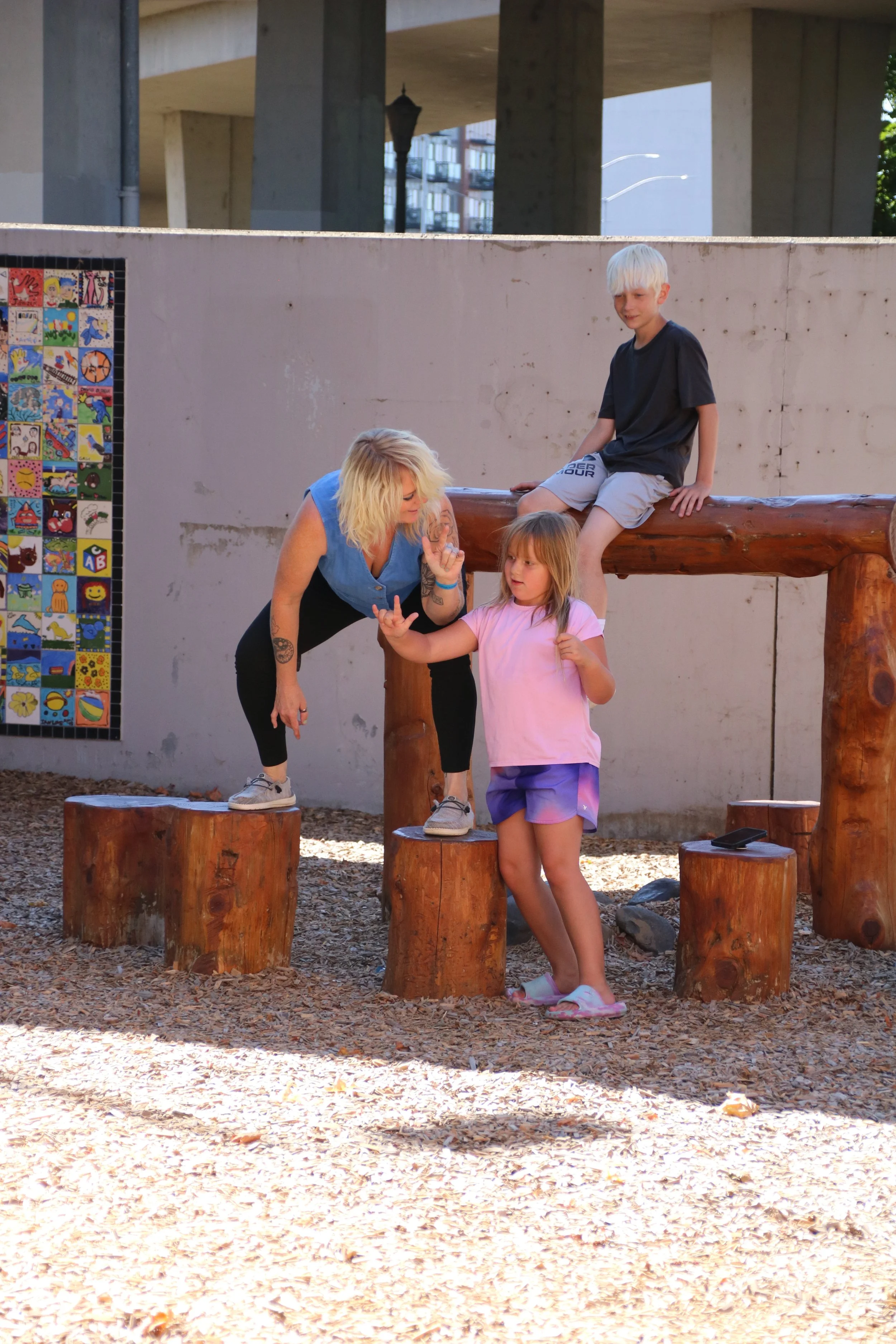 A woman and two children playing on wooden stumps in a playground.