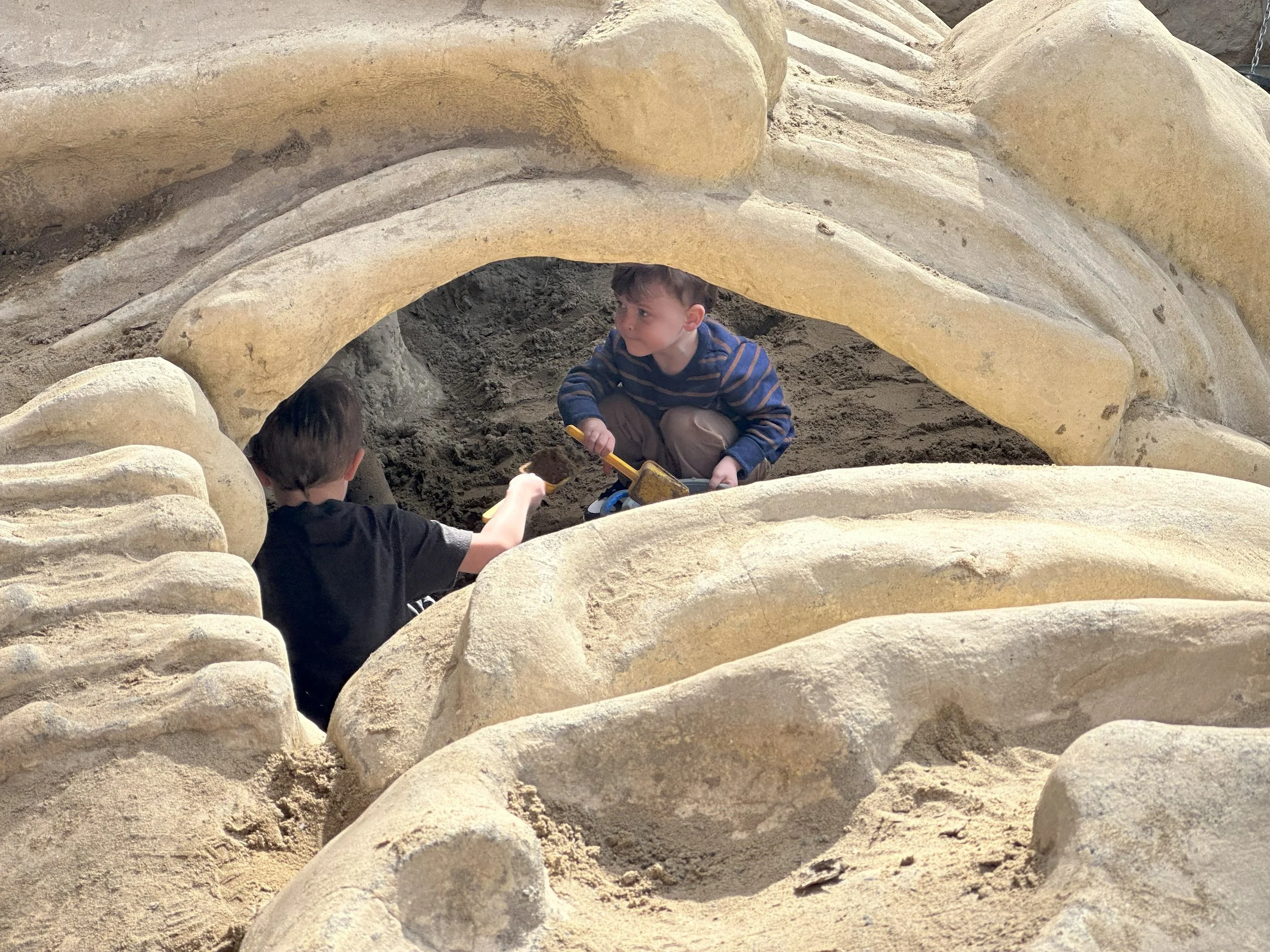 Two children playing in a sandbox under a castle-like sand structure with arches and walls.