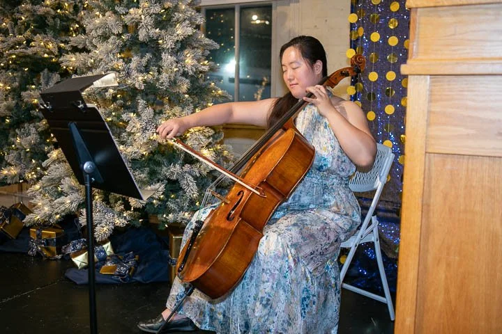 A woman playing a cello while seated next to a decorated Christmas tree, with presents underneath.