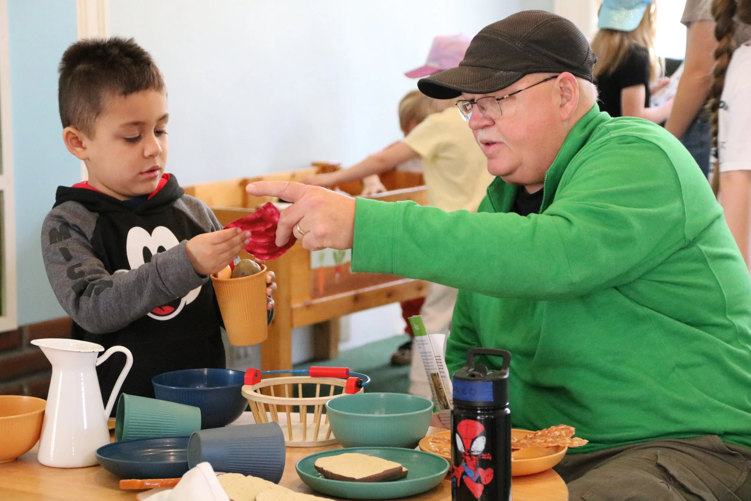 A man in a green jacket and black cap is pointing at a toddler boy holding a yellow cup and a red object, in a room with other children. The table has bowls, a pitcher, a sandwich, and a water bottle with a Spiderman logo.