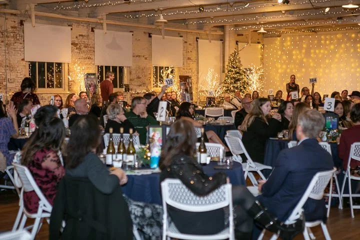Indoor holiday party with decorated Christmas tree, string lights, and multiple round tables with guests in festive attire, bottles of beer, and table numbers, in a rustic brick-walled venue.