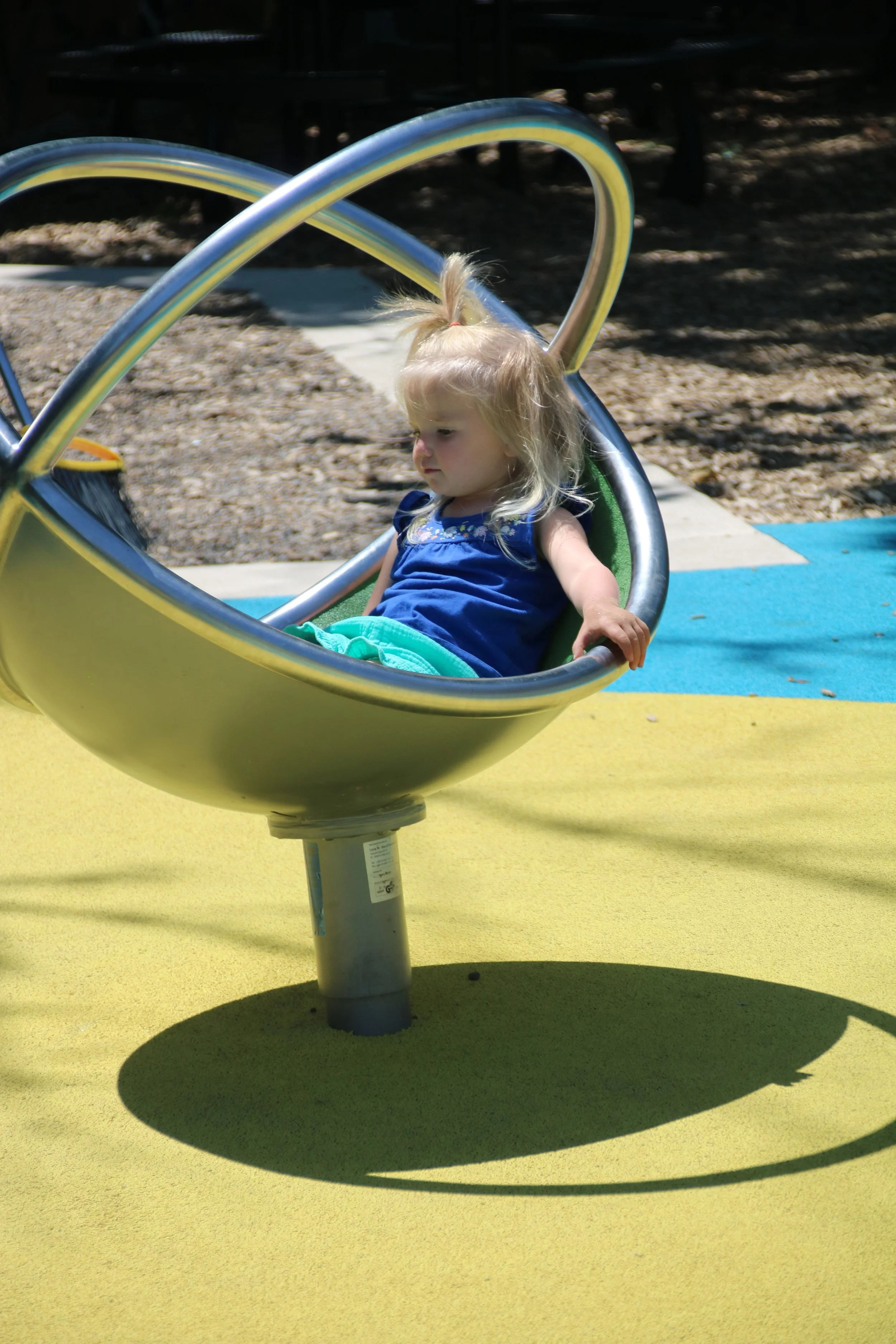 A young girl with blonde hair sitting alone on a playground roundabout or merry-go-round.