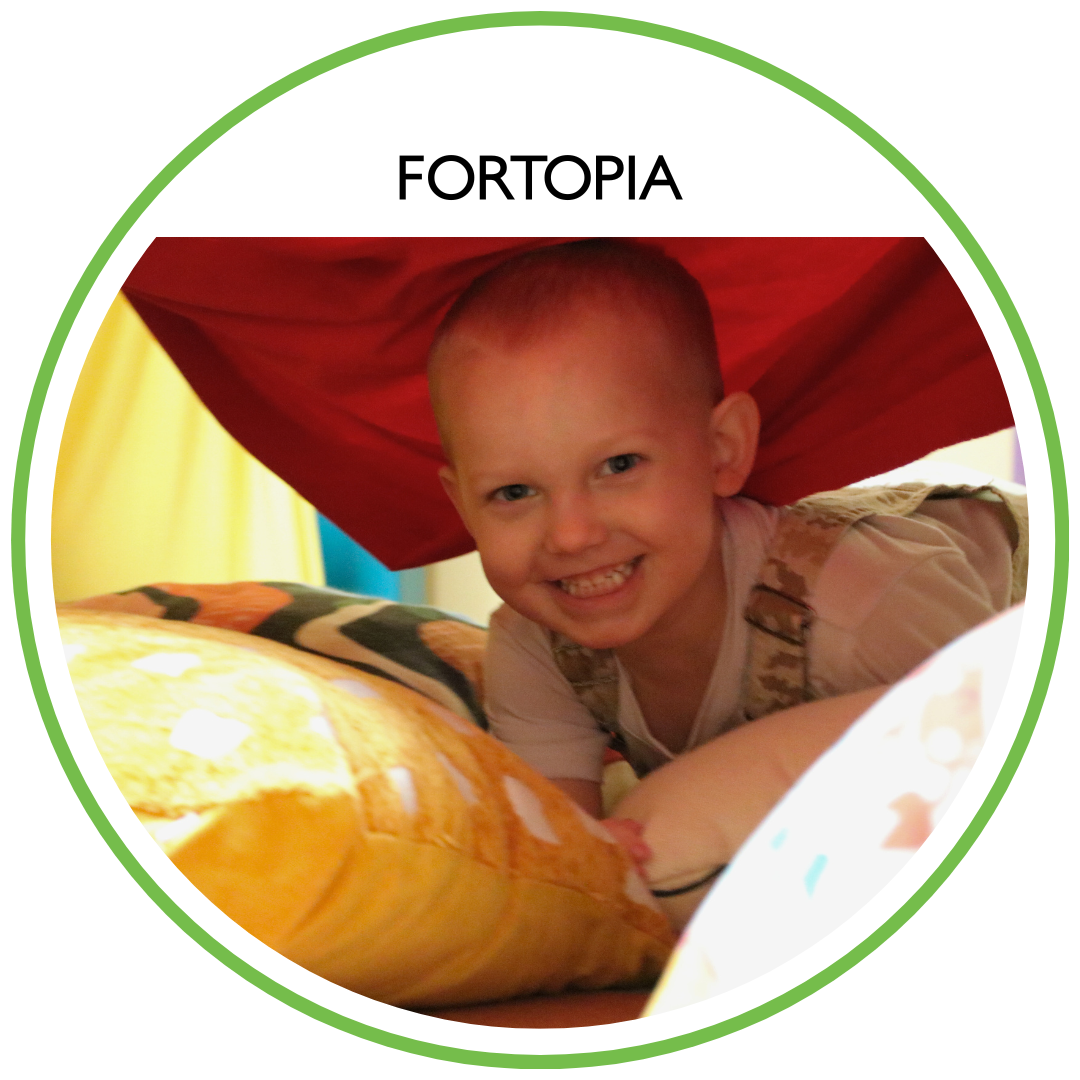 Child smiling and peeking out from under a red blanket or parachute inside a fort cushion play area.