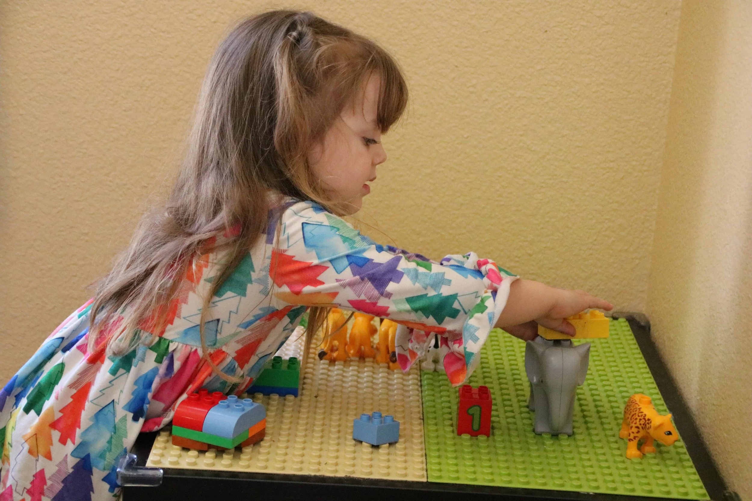 A young girl playing with colorful toy building blocks and animal figures on a LEGO-like baseplate, standing in a room with yellow walls.