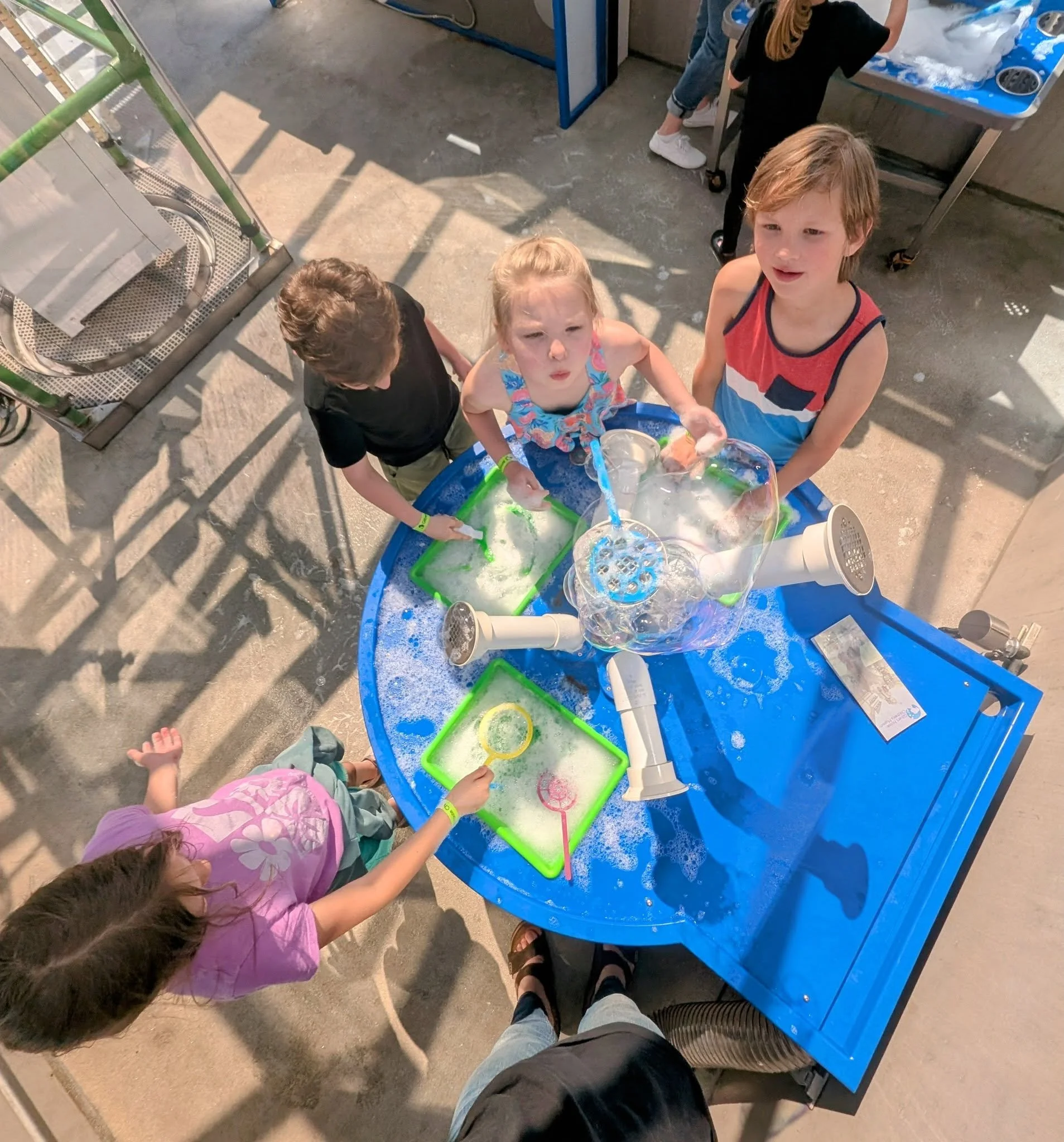 Children playing with a bubble-making science experiment at a blue outdoor table on a sunny day.