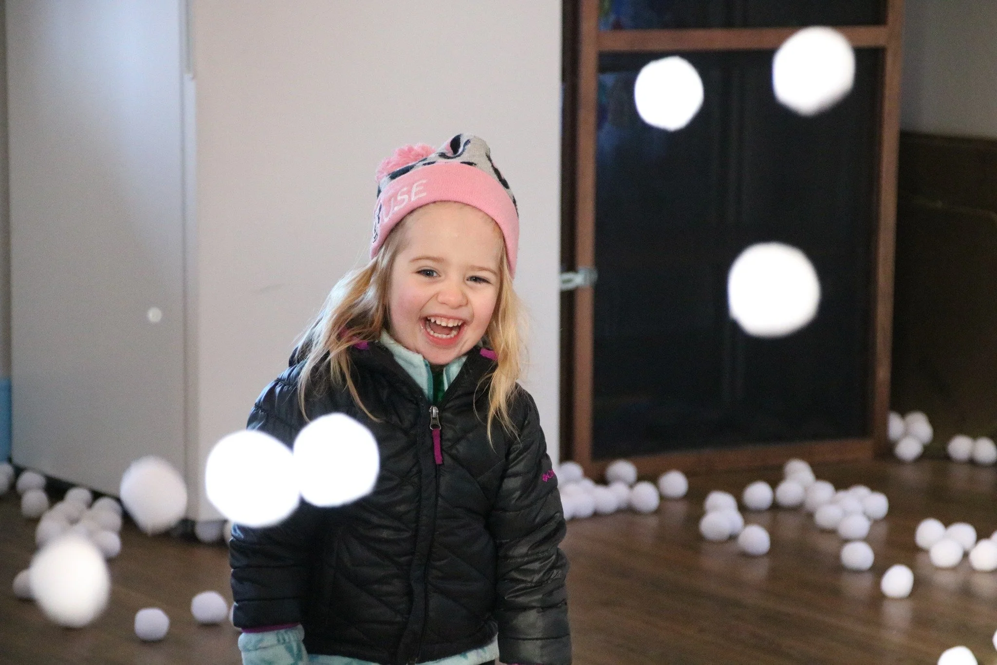 Young girl laughing inside a room with white balls scattered on the floor, wearing a black jacket and pink beanie.