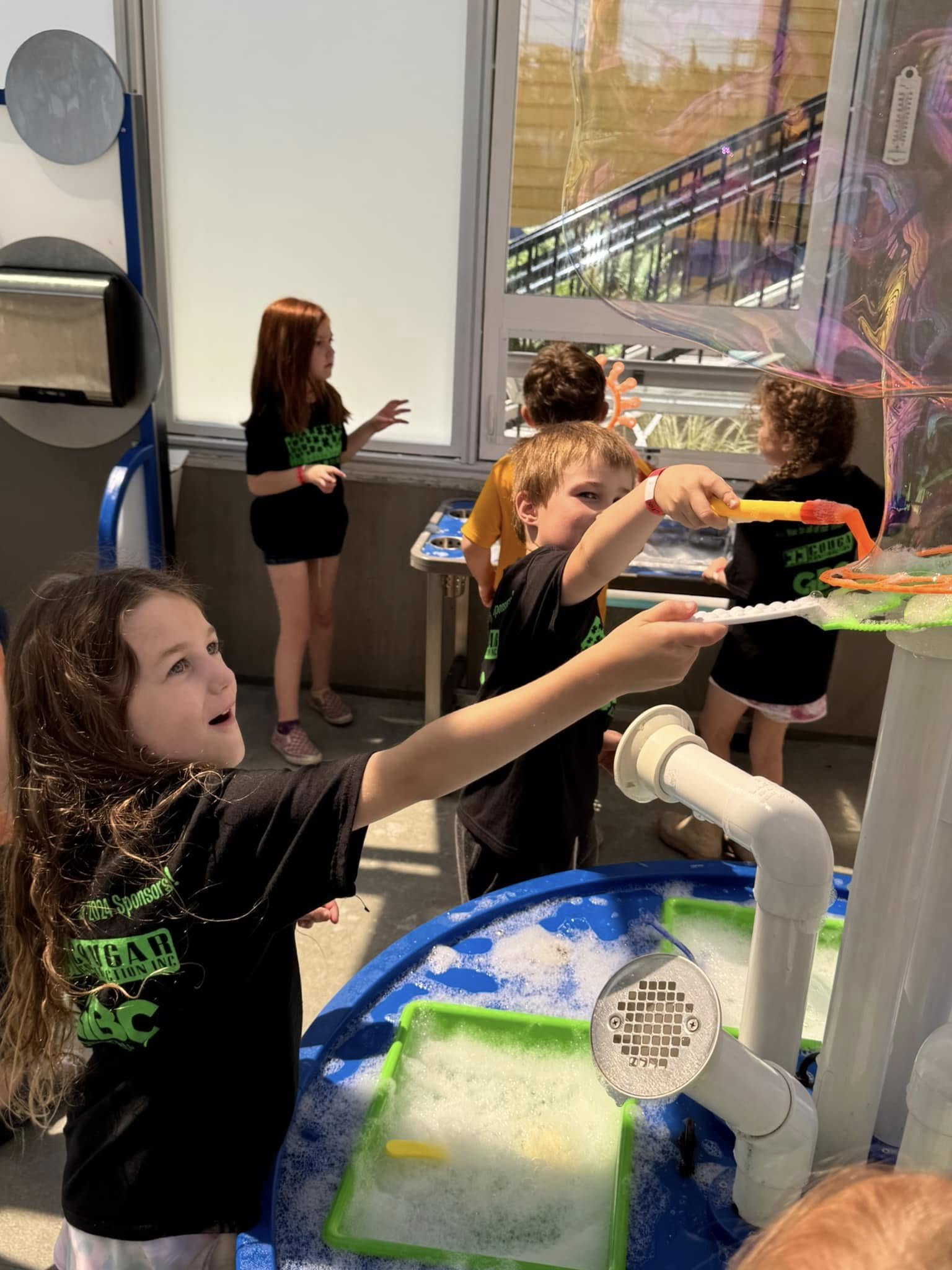 Children engaging in a hands-on science activity at a bubble experiment station, with children wearing black and colorful shirts, and a window in the background showing an outdoor area.
