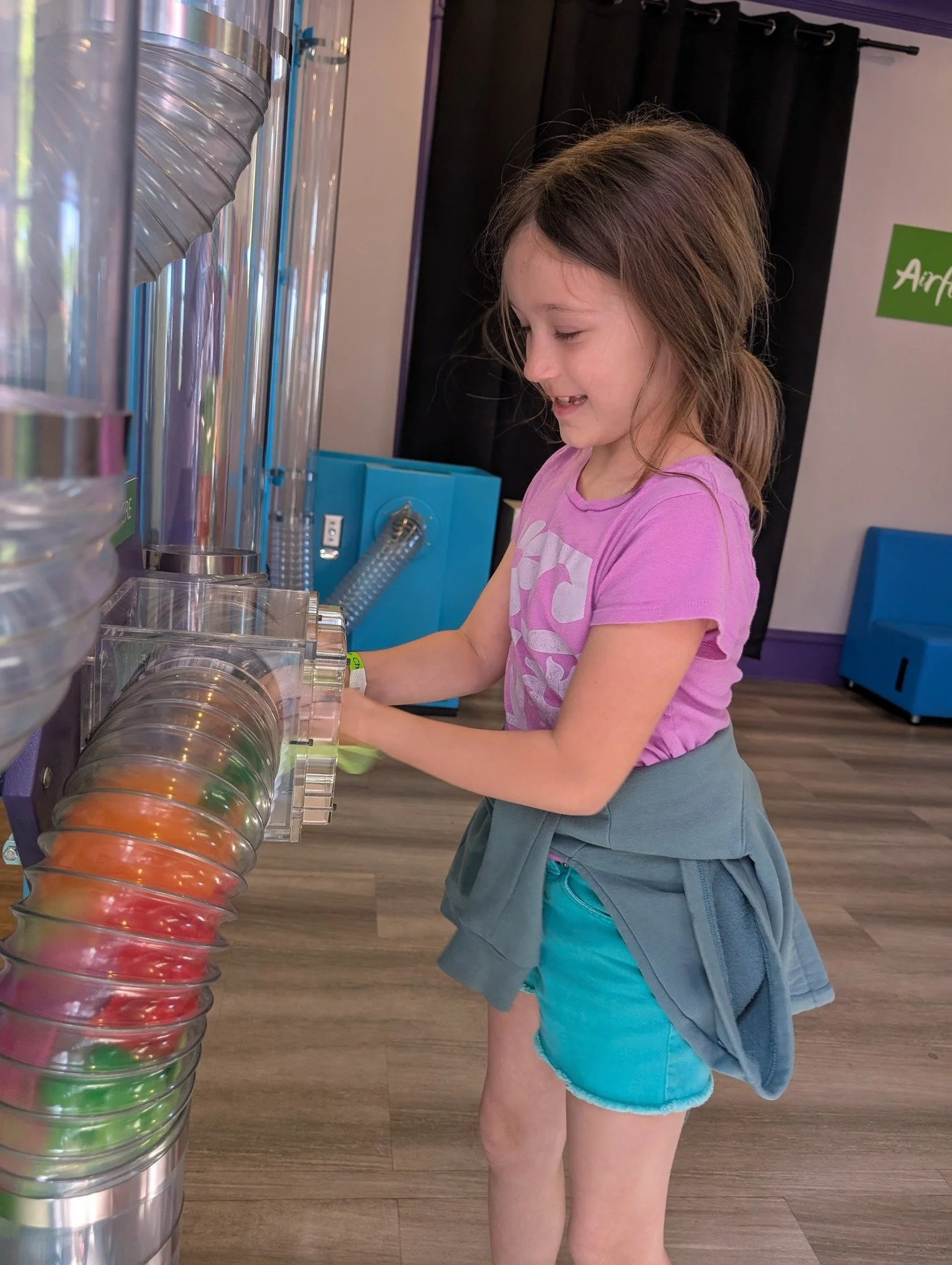 A young girl wearing a pink t-shirt and turquoise shorts interacting with a transparent arcade game or vending machine that dispenses colorful plastic balls.