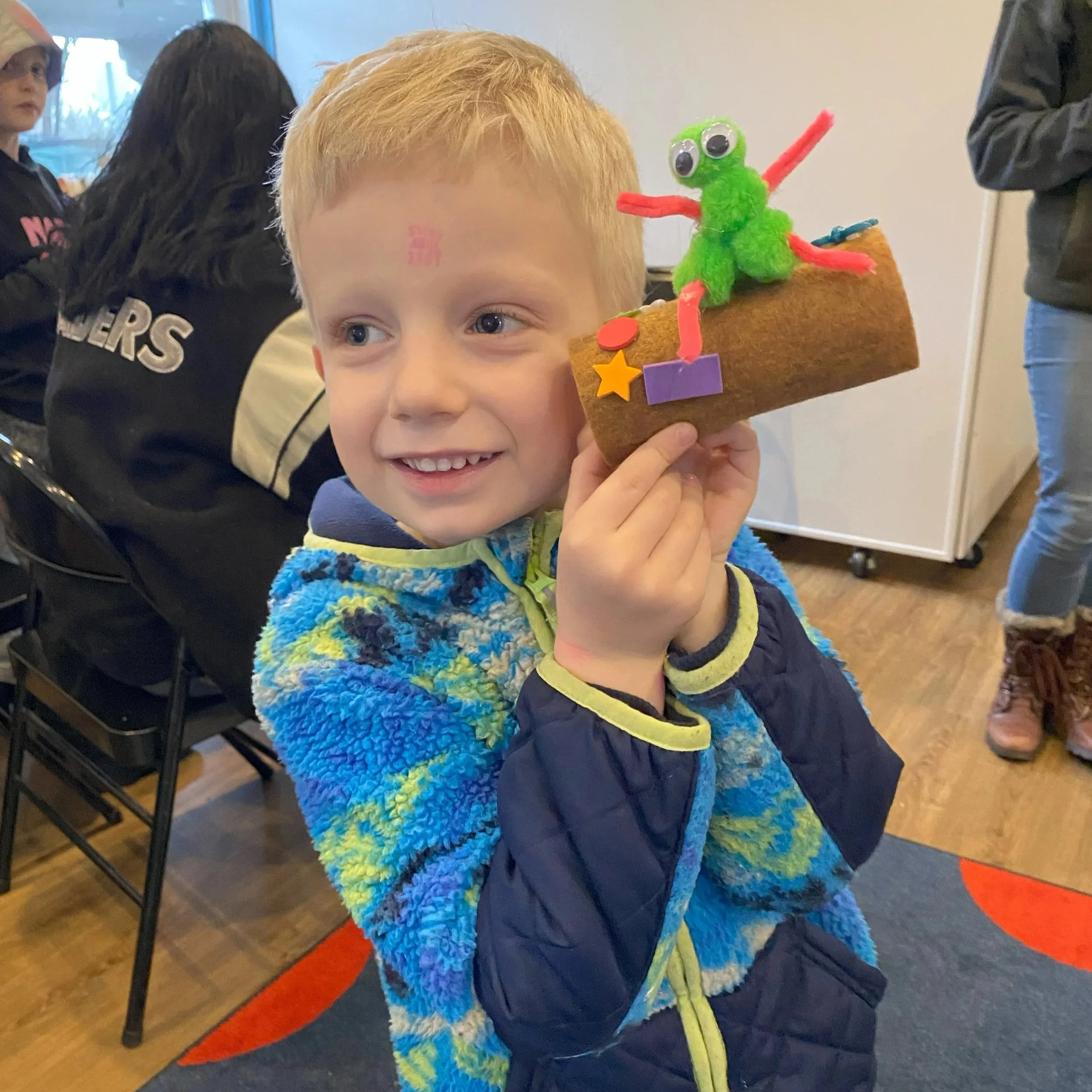 A young boy with blond hair smiling and holding a felt craft of a green alien with googly eyes, sitting on a brown log with colorful decorations. He is wearing a blue and green fuzzy jacket.