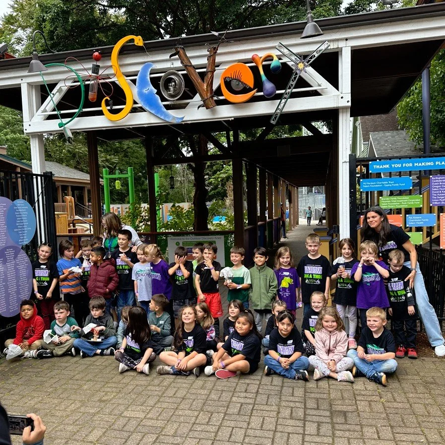 A large group of elementary students sitting and standing in front of the Outdoor Discovery Area entrance gate, which features a colorful "Discover" sign made of artistic letters and shapes.