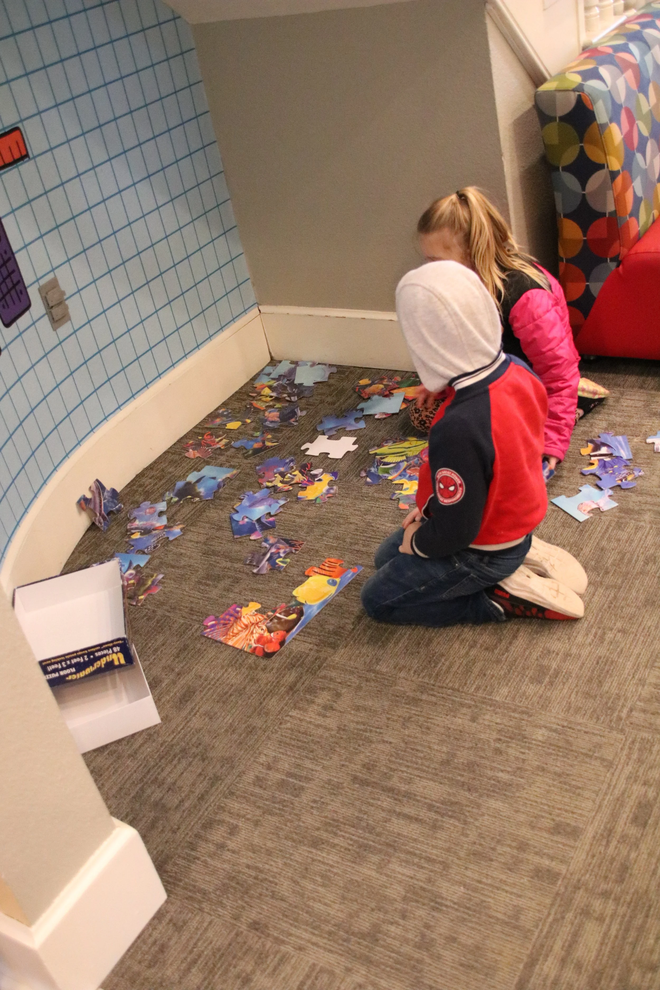 Two children kneeling on the carpeted floor, working on assembling a colorful jigsaw puzzle with many pieces scattered around.