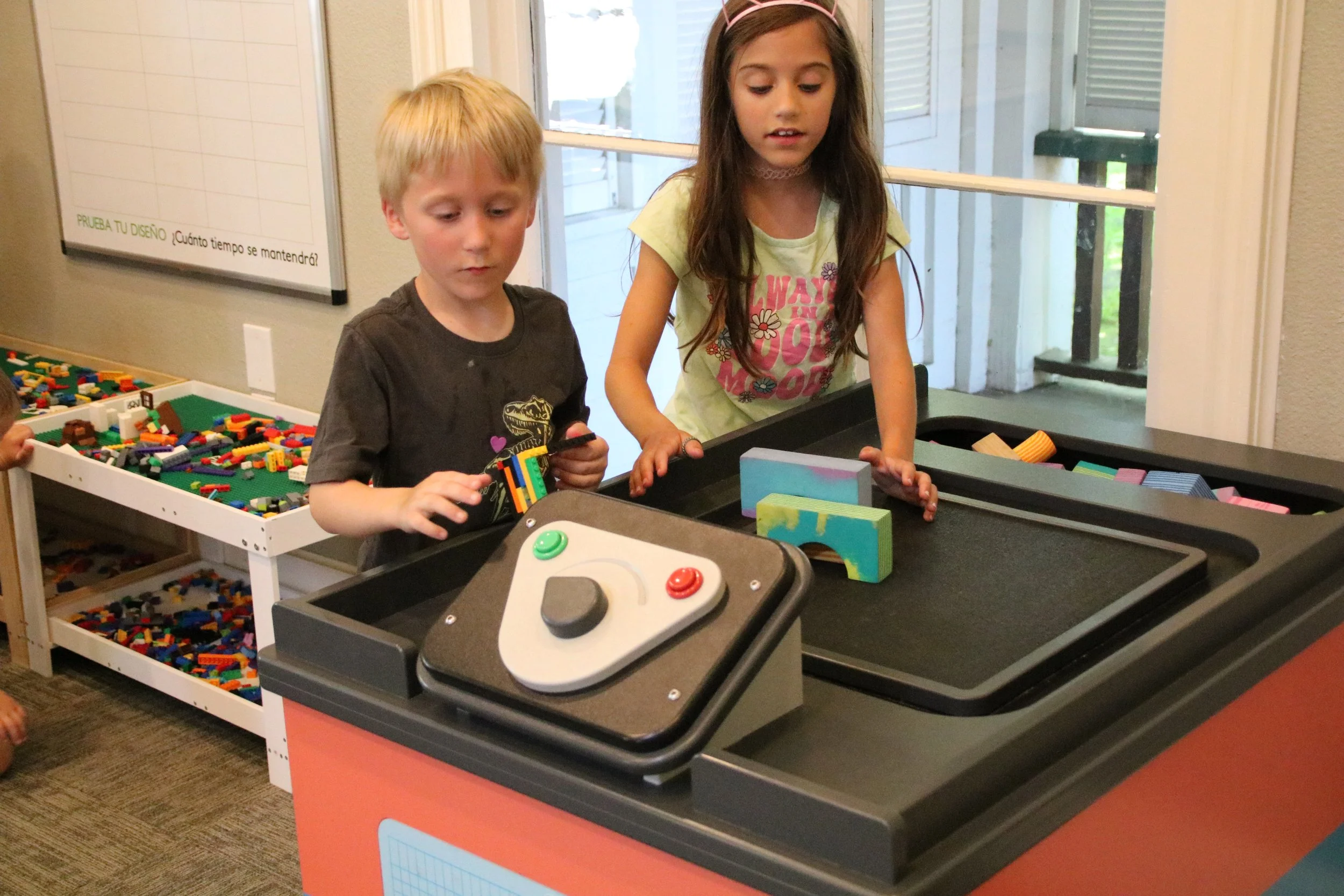 Two children playing with colorful building blocks at a black table in an indoor play area, with a white shelving filled with LEGO bricks in the background.