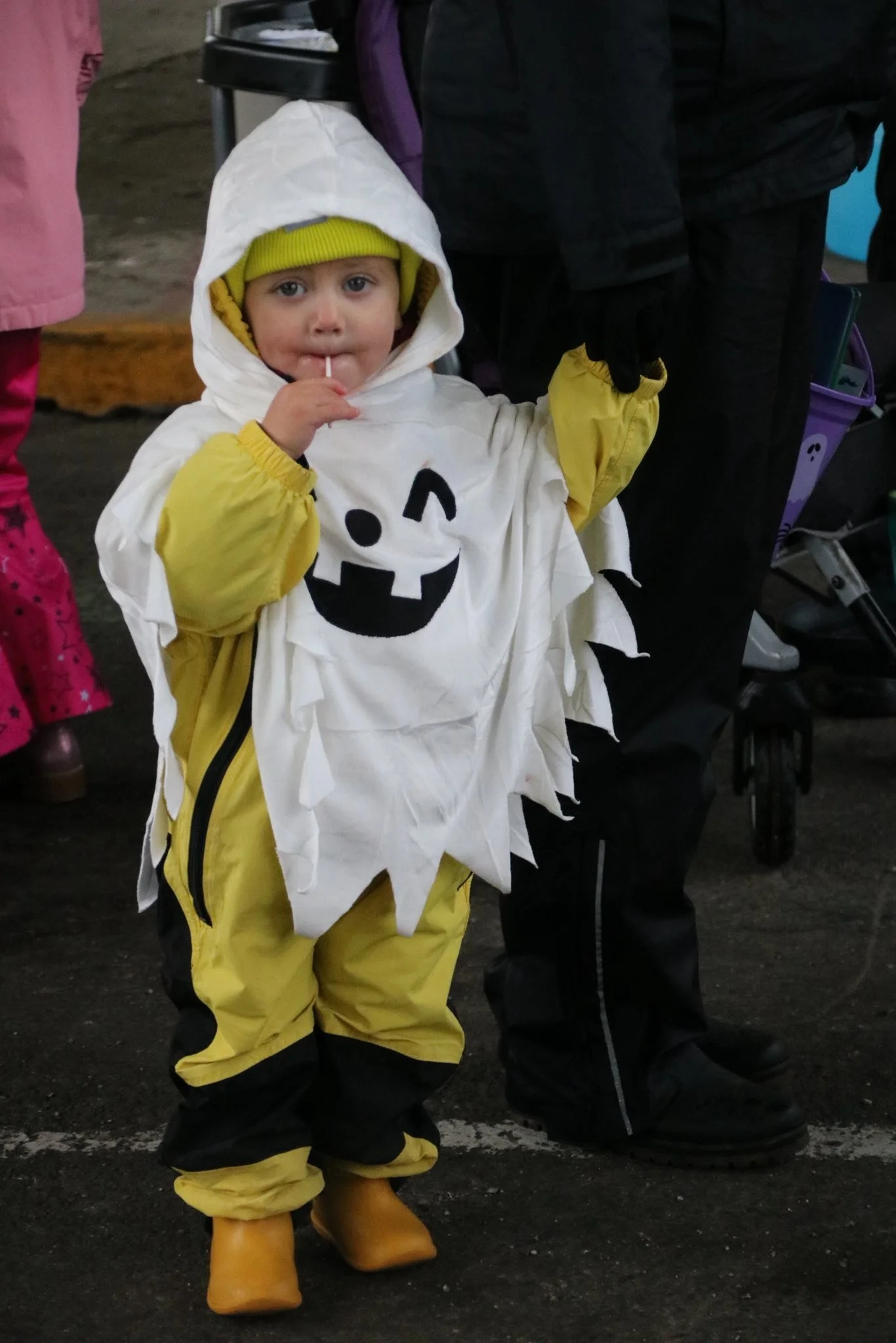 Child dressed in a Halloween costume resembling a ghost or spirit with a pumpkin face, yellow raincoat, yellow hat, and yellow boots, standing in a crowd.