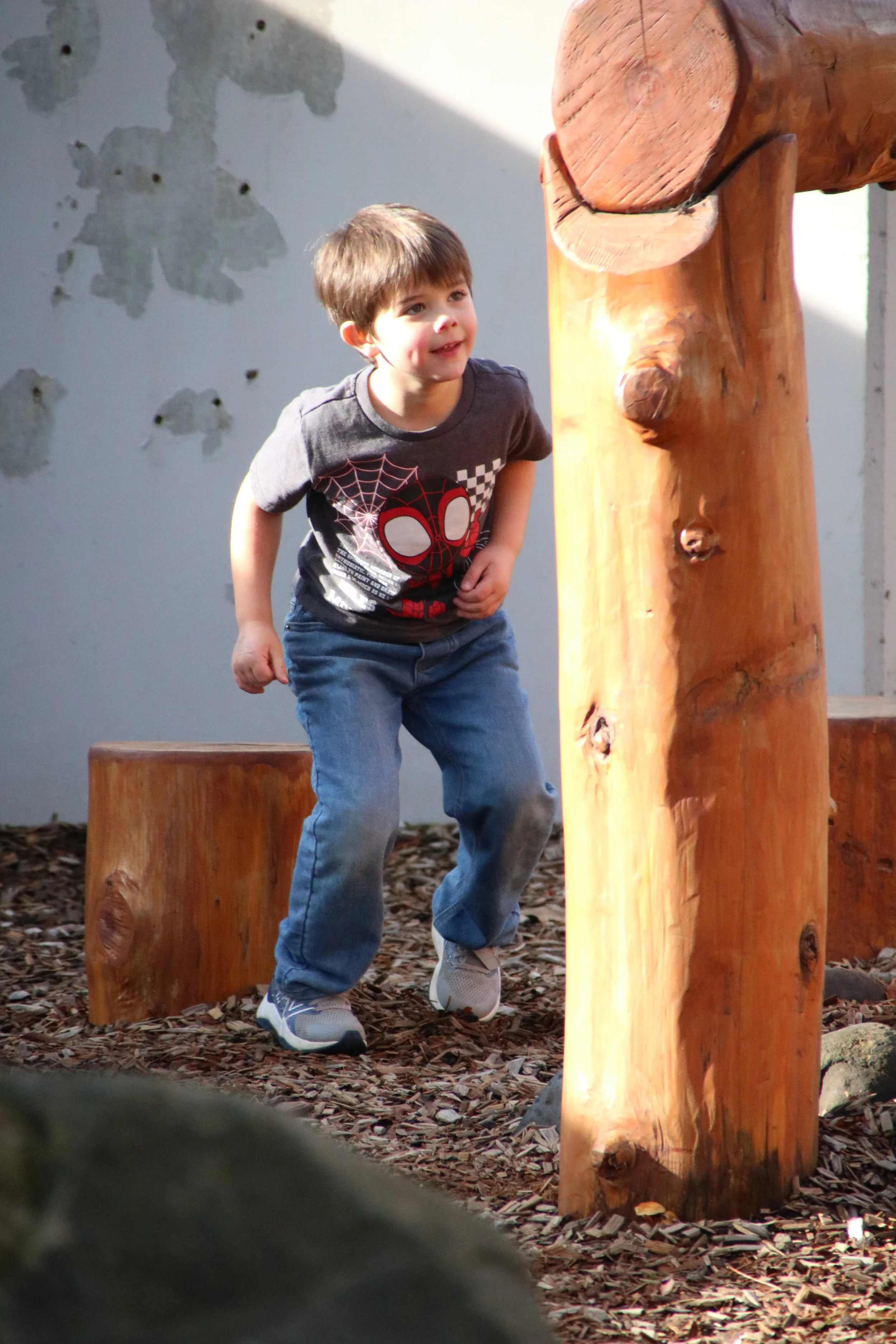 A young boy wearing a Spider-Man t-shirt and jeans is jumping on a wooden outdoor play structure.