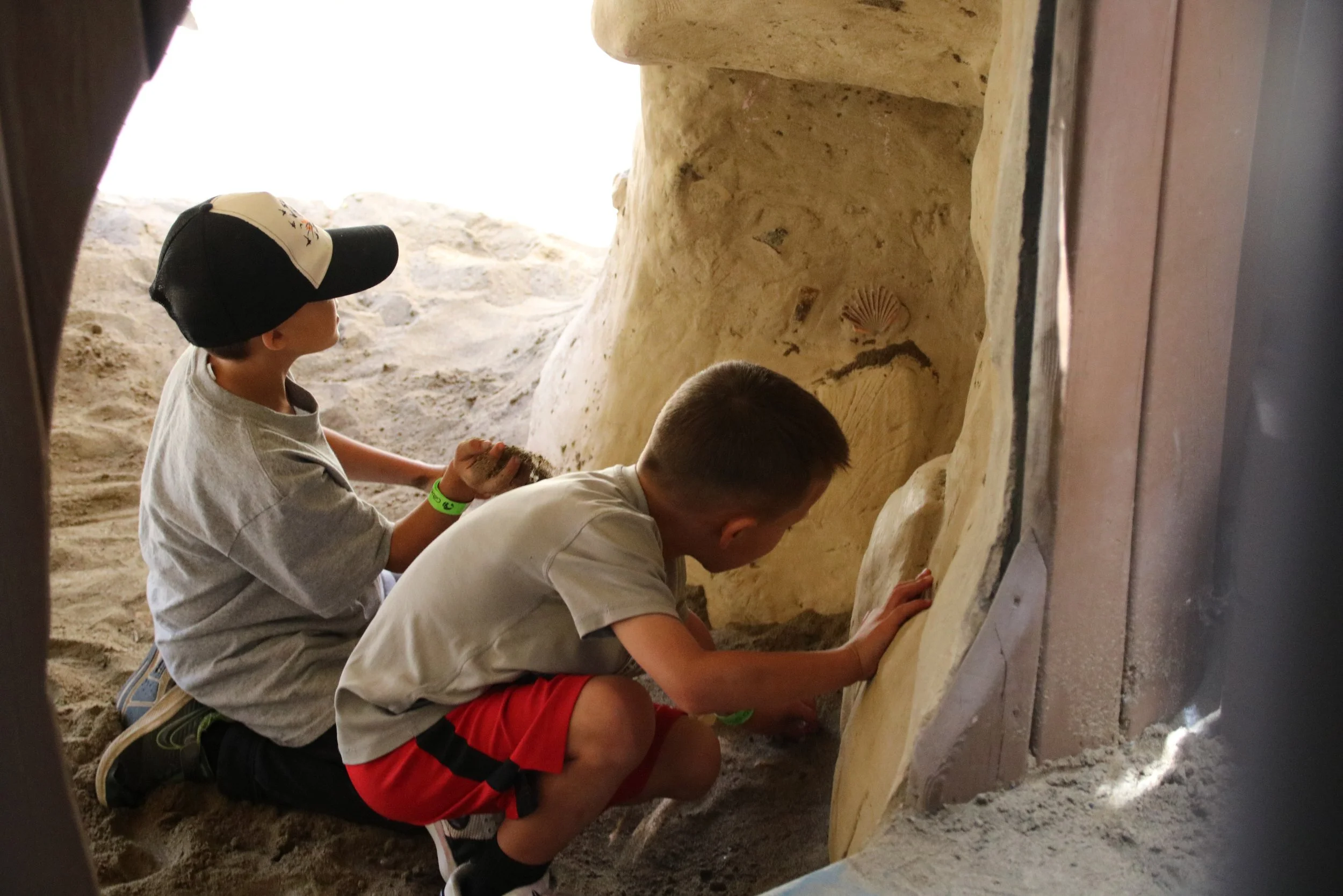 Two boys crouching behind a wall, playing in a sandy area, with one touching the sand and the other examining a shell on the wall.