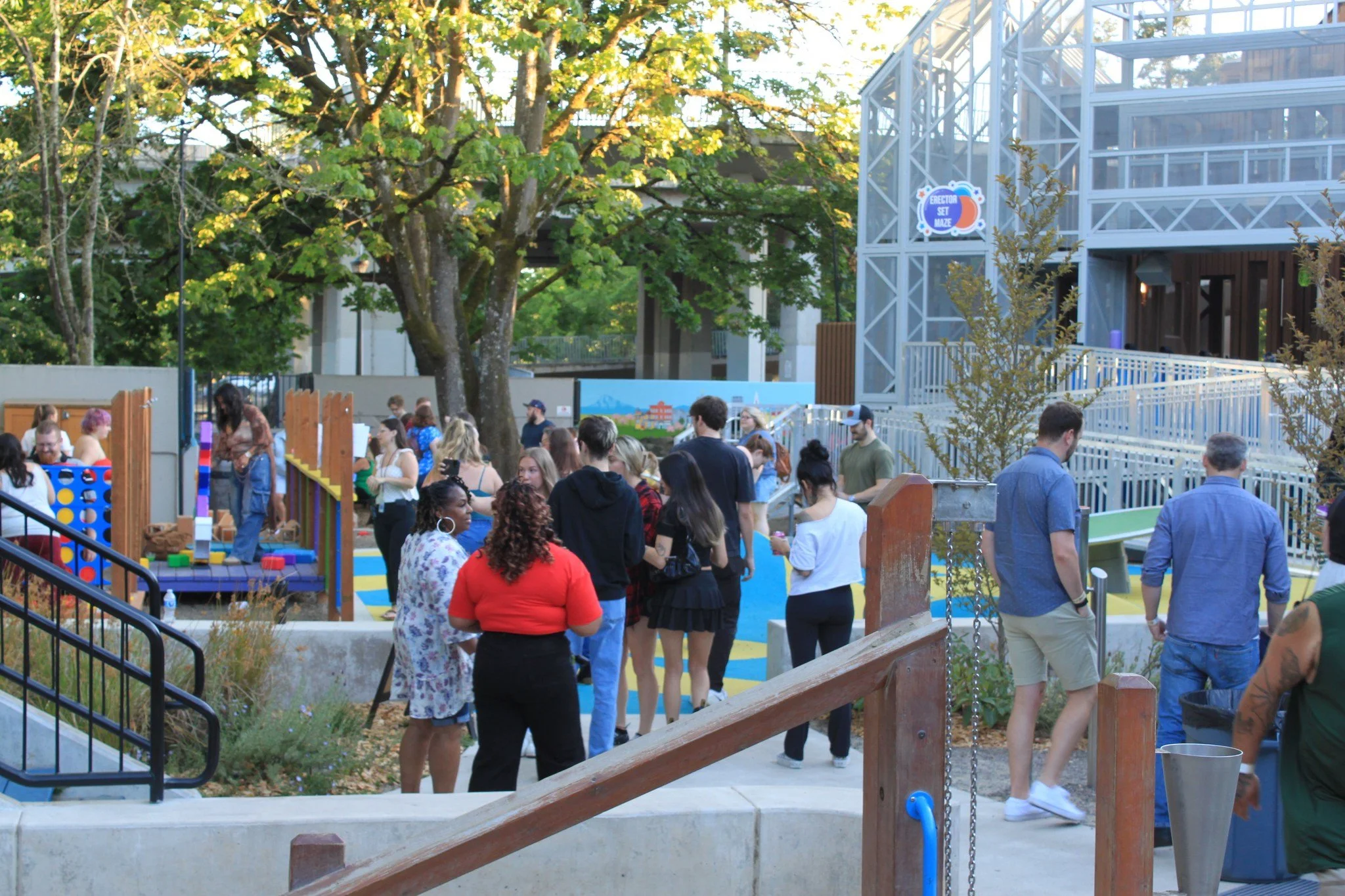 Group of people gathered at a colorful outdoor playground area with trees and a building in the background.