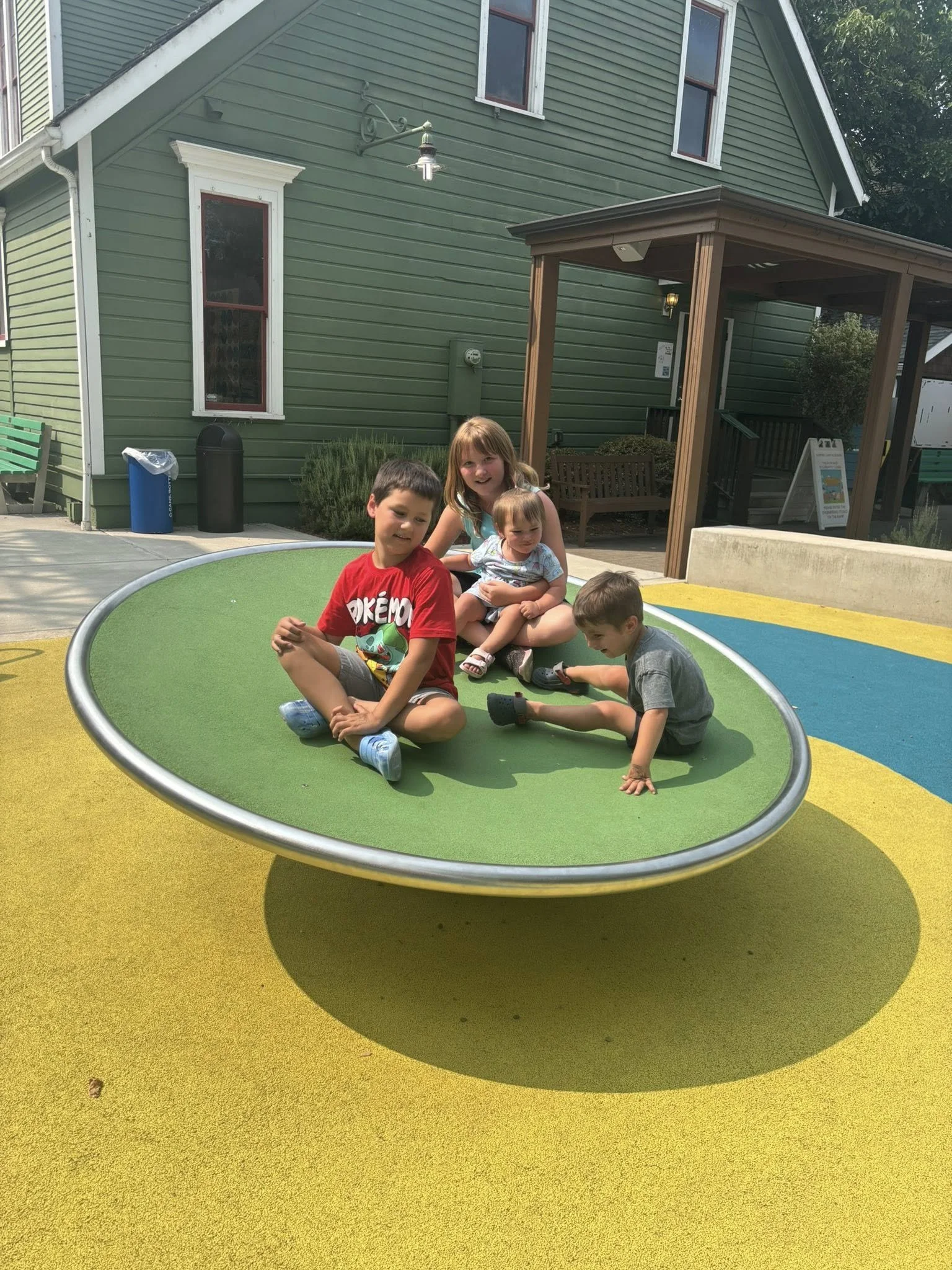 Four children sitting on a large green circular play surface in a colorful playground area, with a green building and a wooden pavilion in the background.