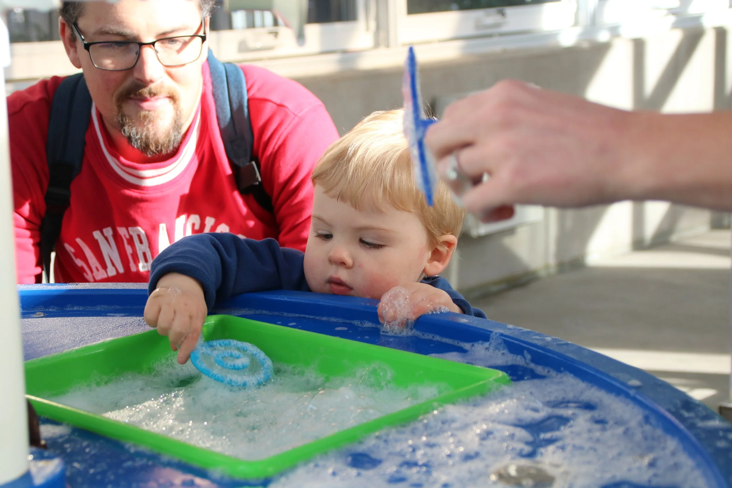 A young boy with blonde hair leaning on a blue water table with bubbles, holding a blue bubble wand, while an adult's hand holds another bubble wand above him. A man with glasses and a red shirt watches nearby.