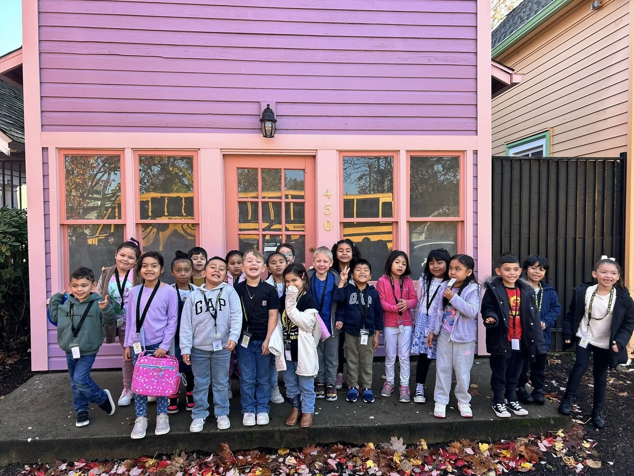 A group of students standing on the porch of a bright purple historic building with pink trim. A yellow school bus is reflected in the front windows.