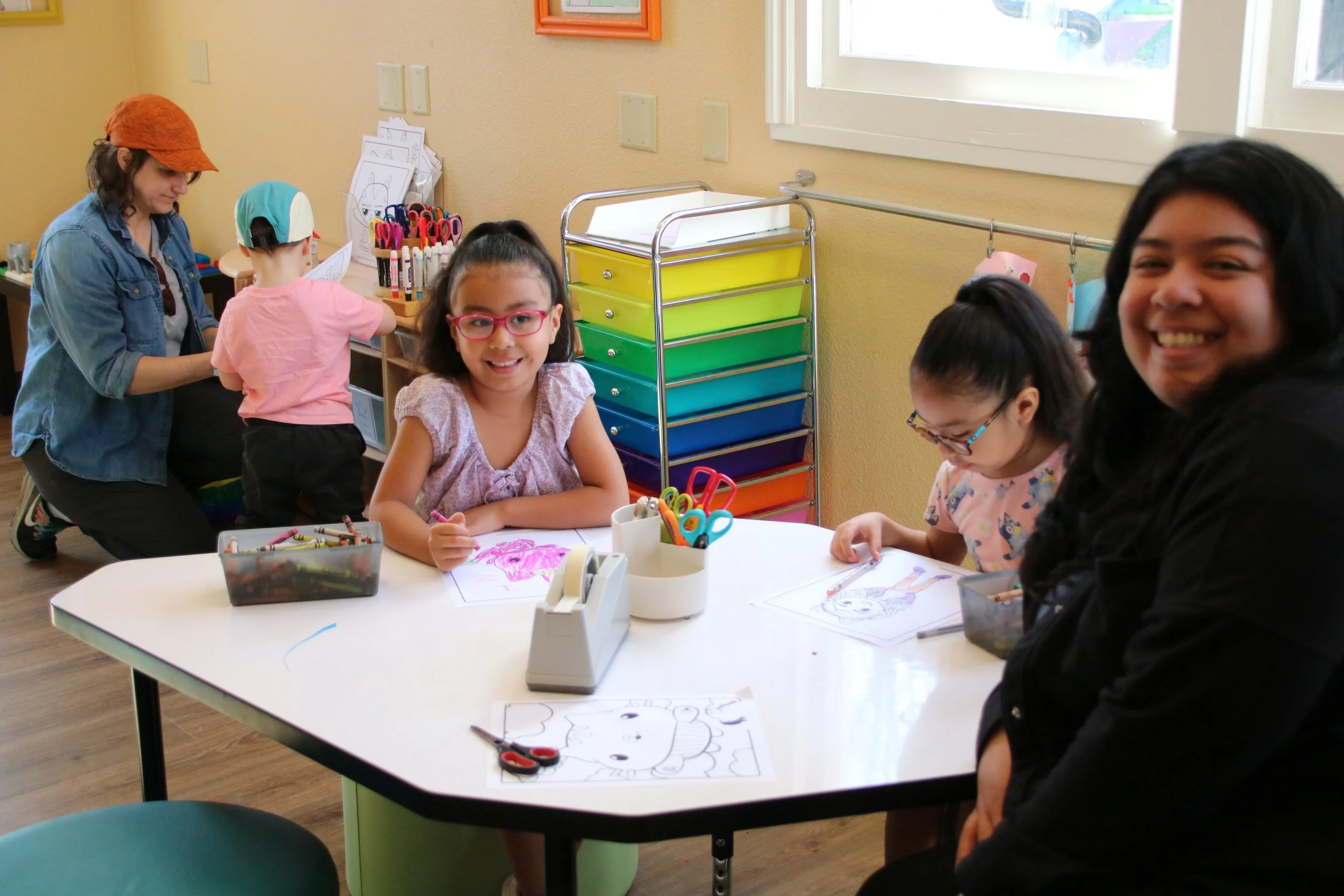 Children and a teacher in a classroom engaging in arts and crafts activities. The children are drawing and coloring at a table with various art supplies.