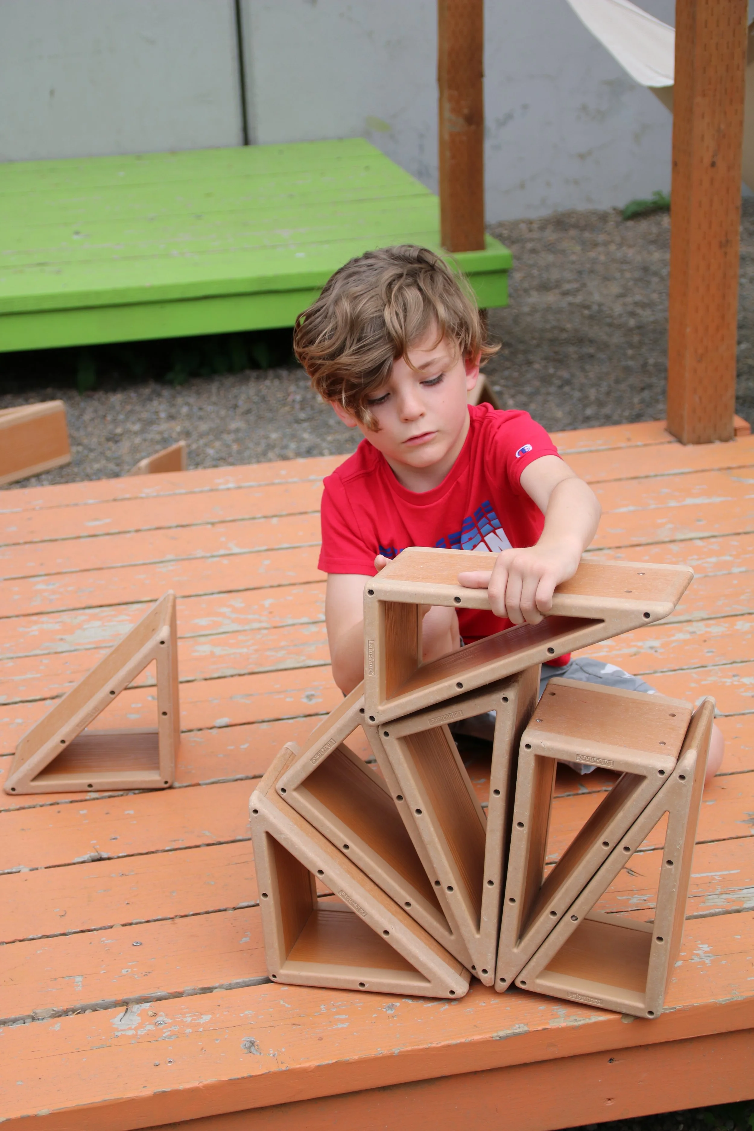 A young boy in a red shirt building a structure with wooden triangle-shaped blocks on an orange wooden deck.