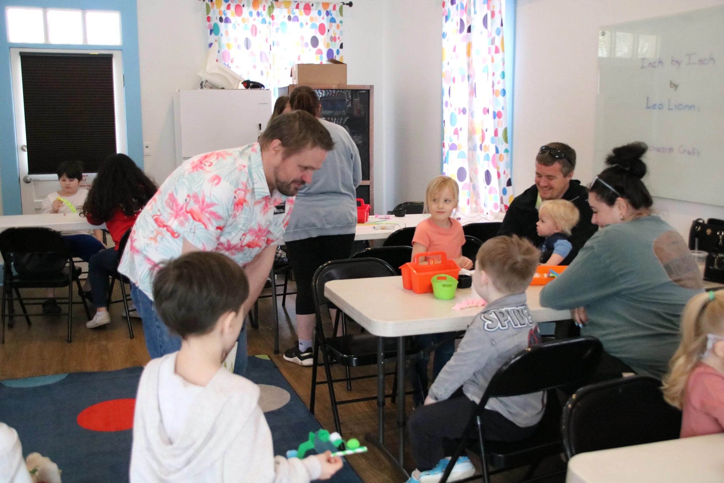 Children and adults in a classroom participating in a crafting activity, with tables, chairs, and colorful curtains in the background.