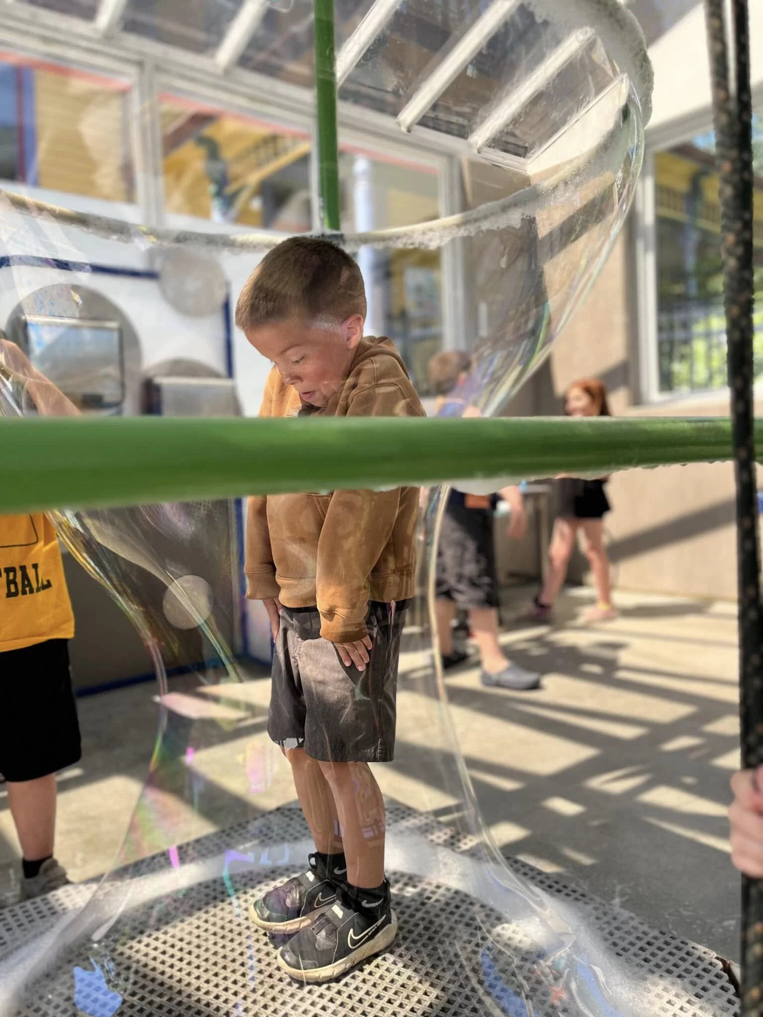 Young boy standing inside a large soap bubble at an outdoor event, with other children and adults in the background.