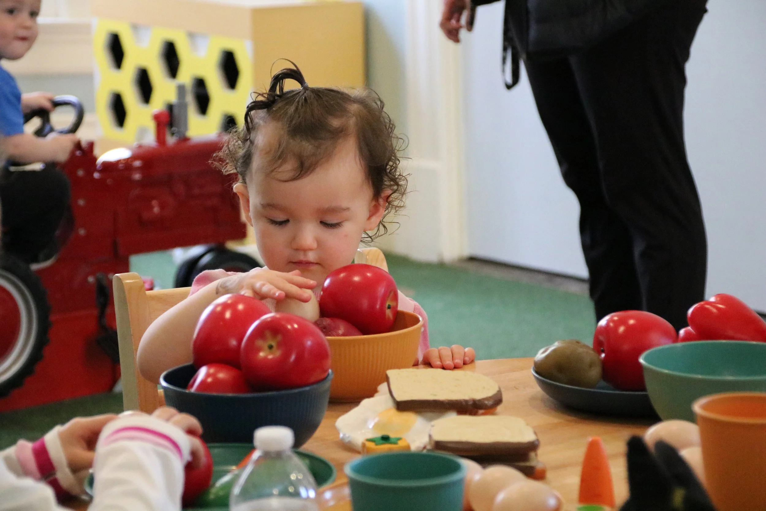 A young girl sitting at a table filled with toy food items, including apples, crackers, and a bottle, in a playroom with a yellow and black honeycomb decoration in the background. She is looking down at the toy apples in front of her.