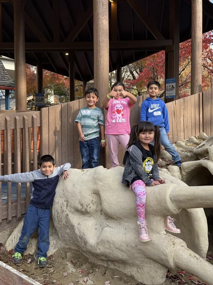 Five children are playing on a stone sculpture that resembles an elephant's head with tusks, outside in a park with autumn trees. Four children are on a wooden platform behind the sculpture, and one girl is sitting on the elephant's snout with her skirt hanging over the side.