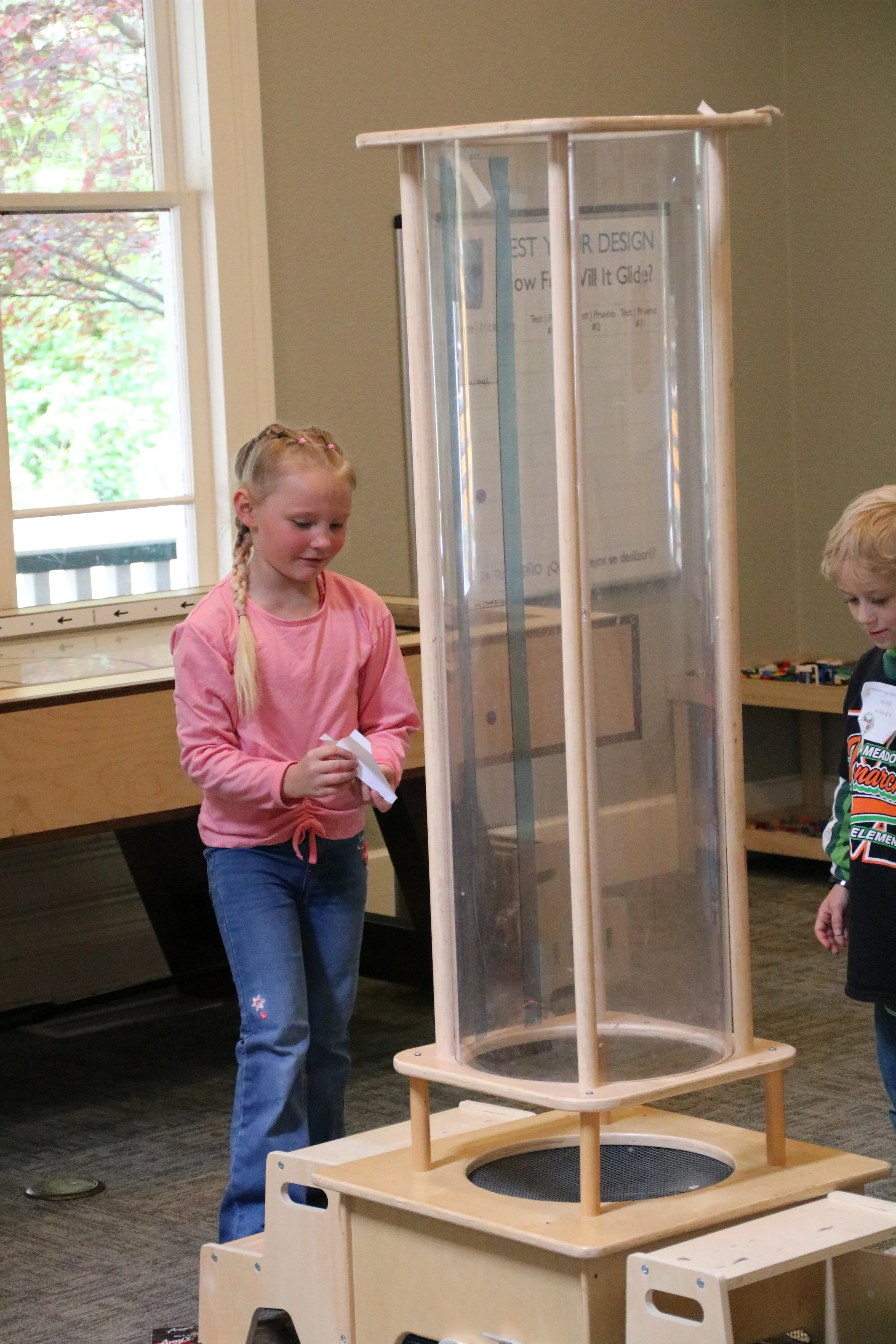 Two children, a girl with blonde braids in a pink shirt and a boy with blonde hair wearing a hockey jersey, interacting with a science or engineering exhibit in a room with a window, a table, and a shelf filled with toys or models.