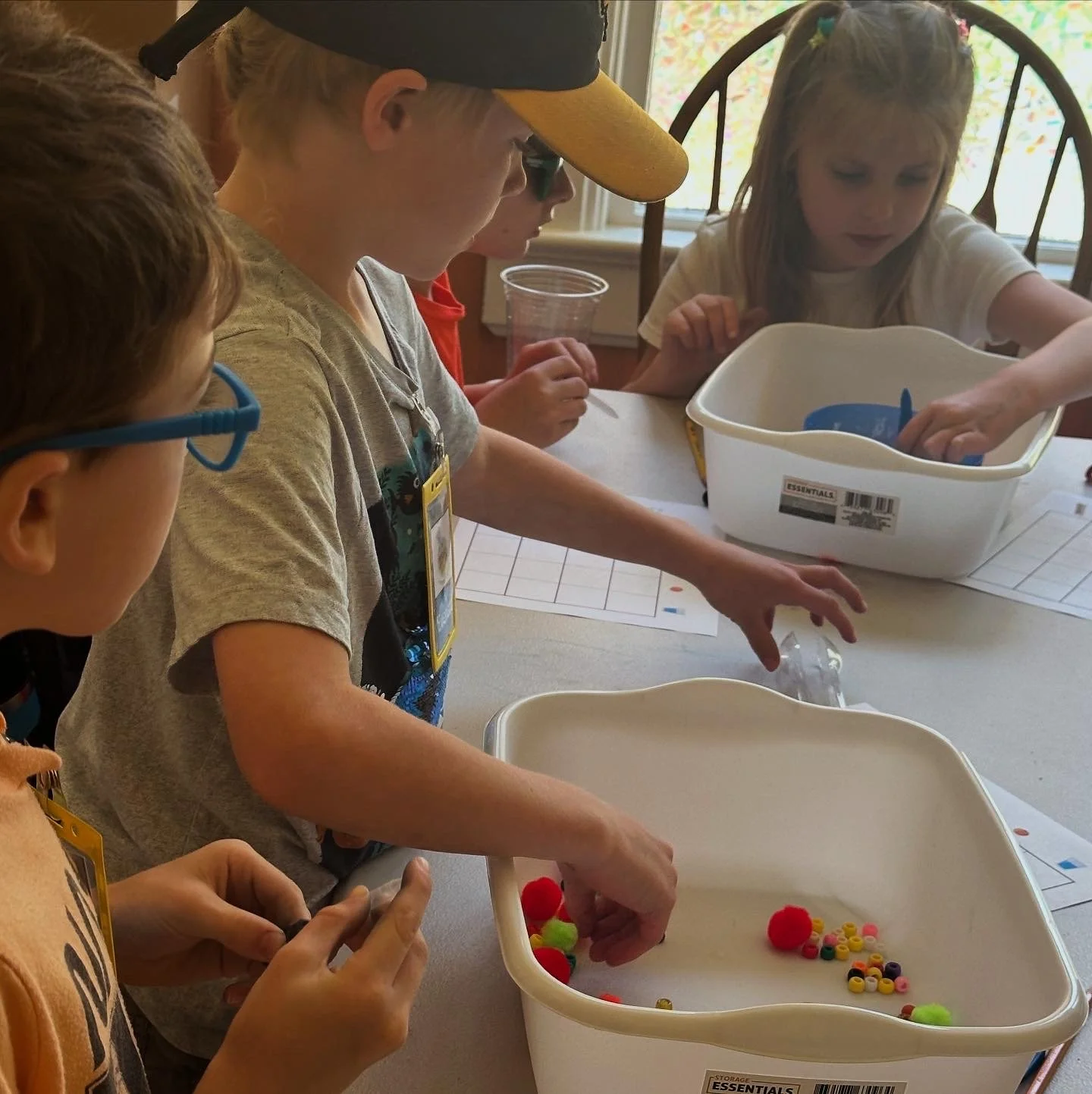 Children gathered around a table working on a craft activity, with colorful pom-poms and beads, involving sorting or assembling objects.