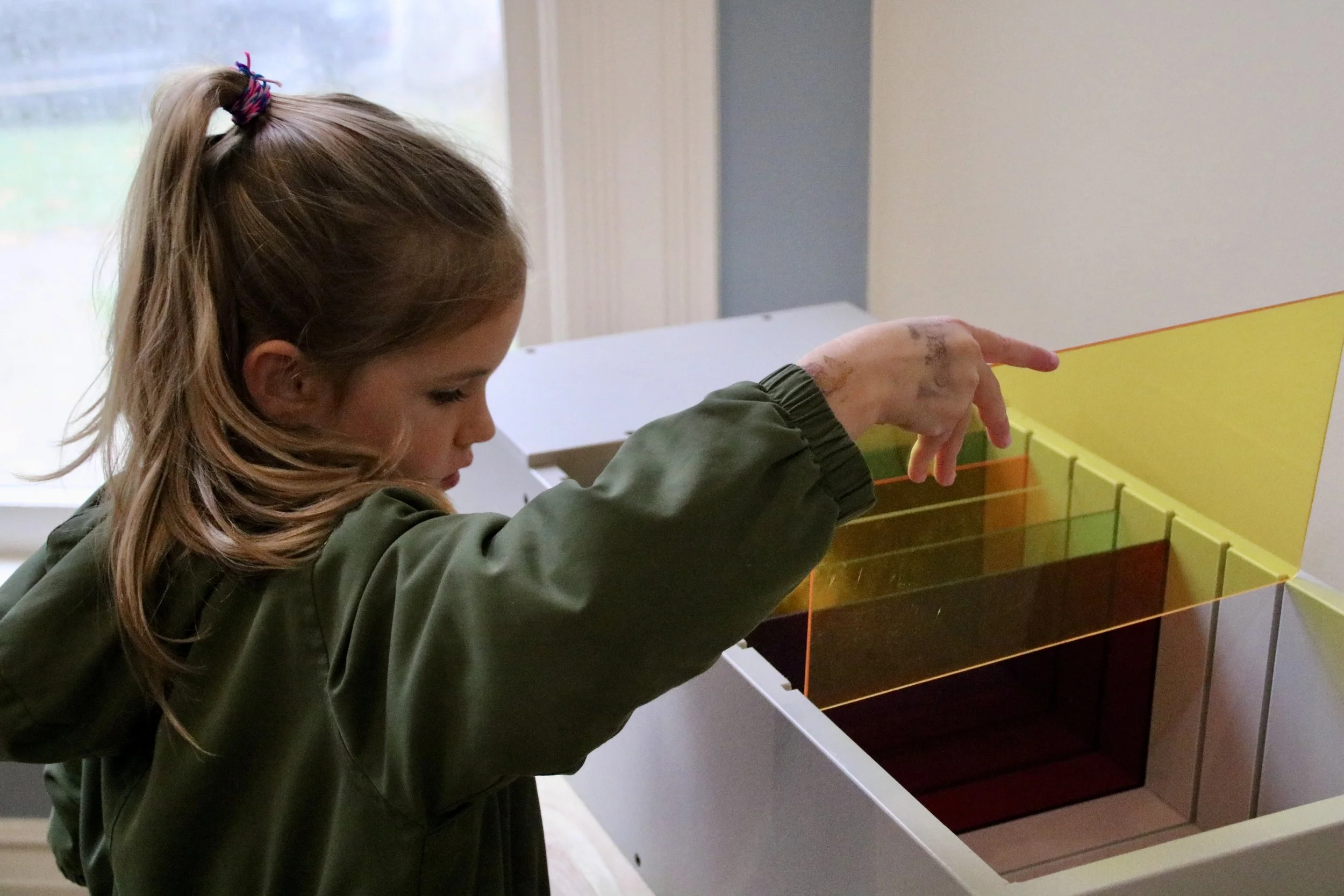 A young girl with long, light brown hair tied with a purple scrunchie is pointing at a rainbow-colored, transparent, tiered display or container.