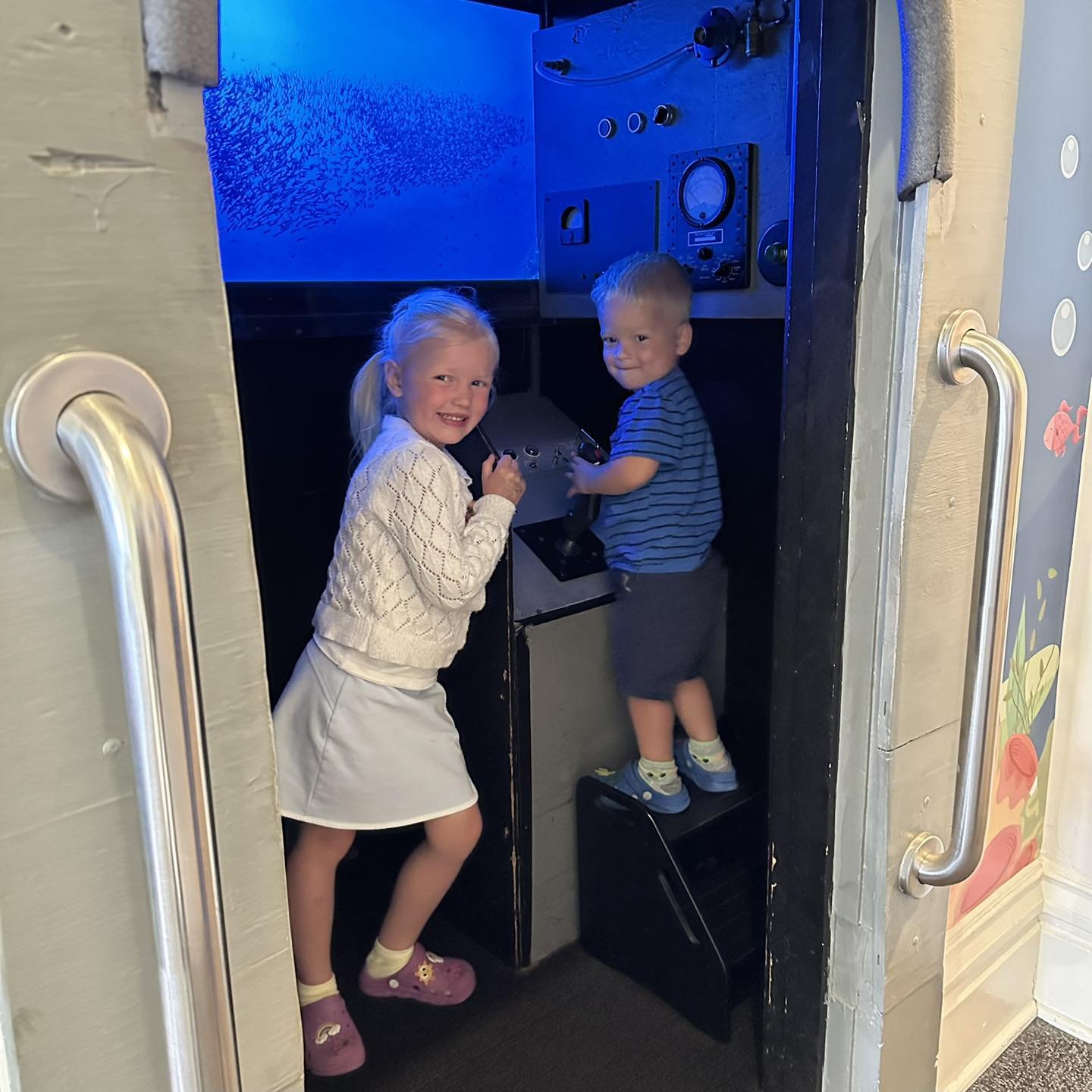 Two young children, a girl and a boy, playing inside a small interactive booth with a control panel, in a brightly lit room with colorful wall decorations.