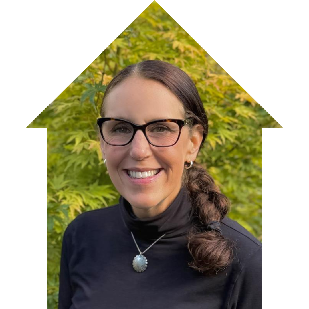 A woman with glasses and a dark top smiling outdoors with green foliage in the background.