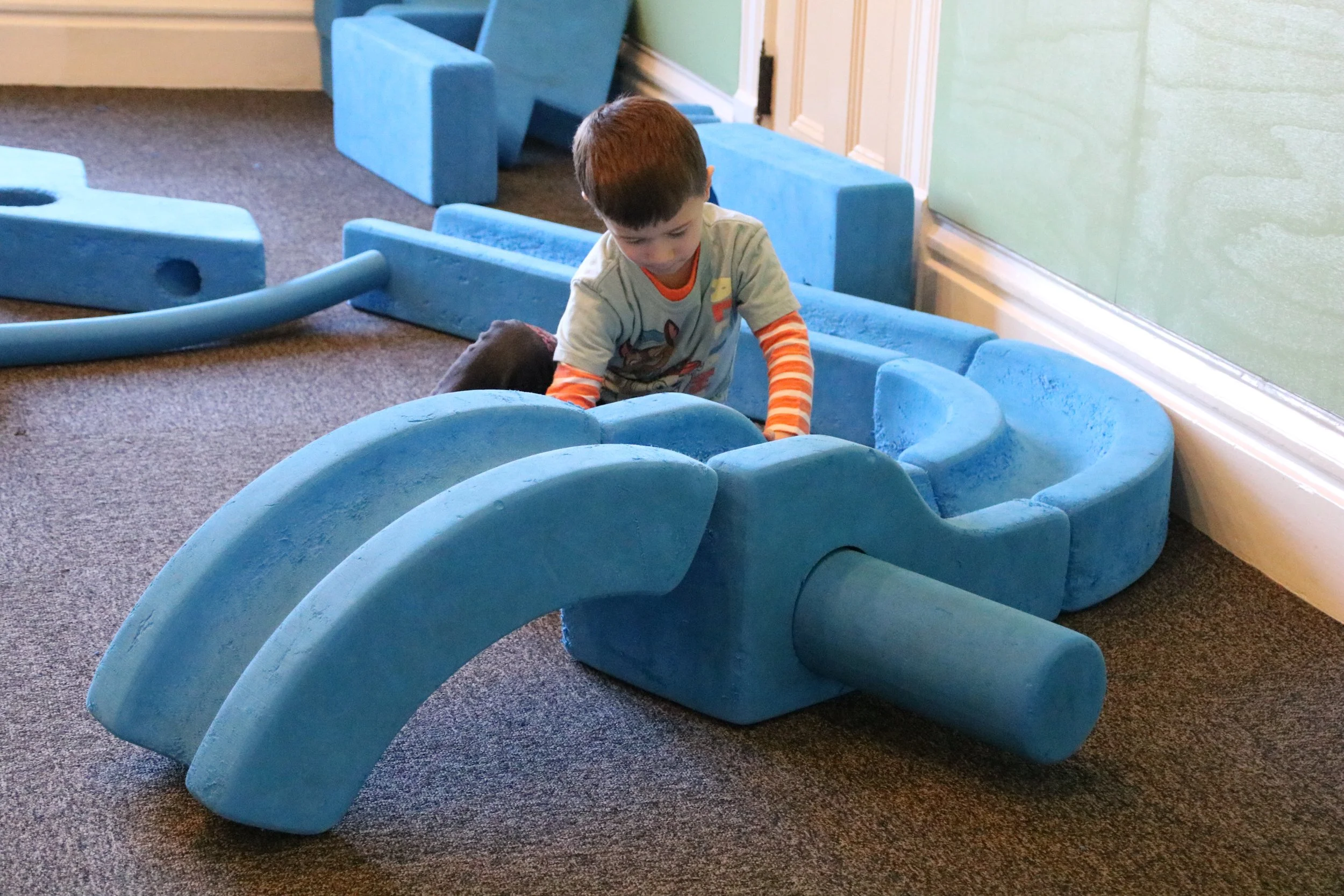A young boy playing with a large, blue, foam toy dinosaur inside a room with carpeted floor and green wall.