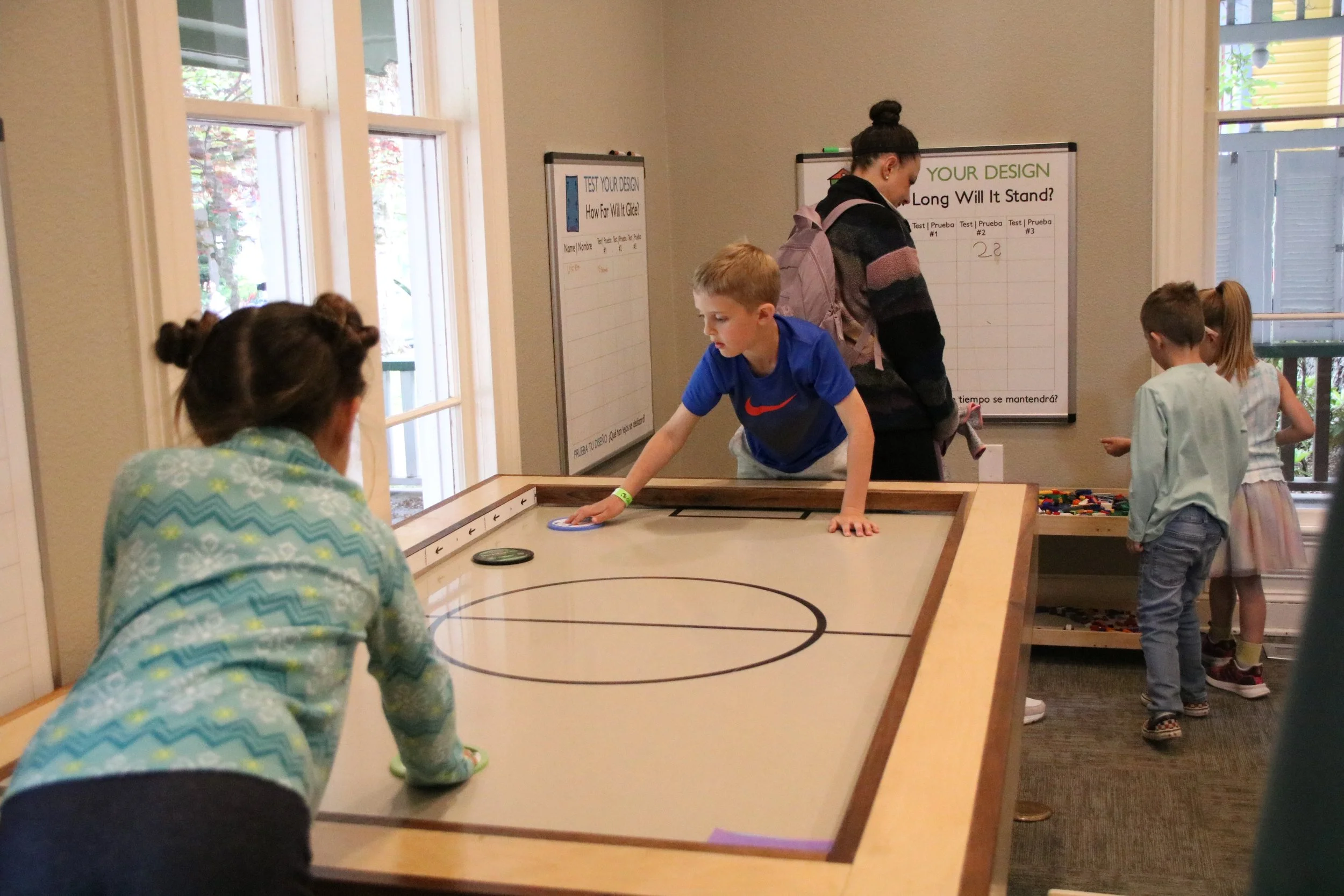 Children playing air hockey in a room with large windows and whiteboards