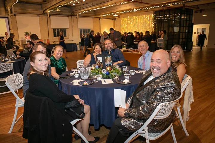 Group of seven people seated at a round table during a celebration or event, with string lights and festive decorations in the background.
