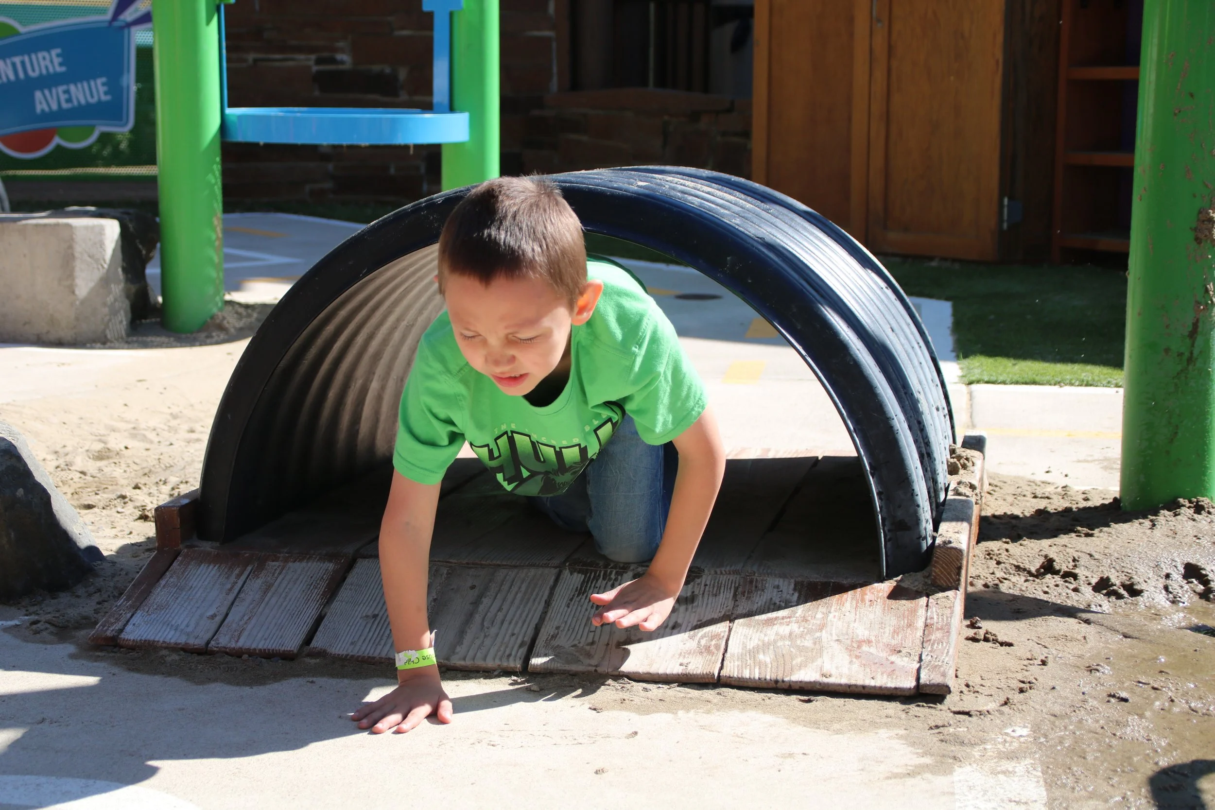 A young boy in a green shirt crawling out of a black tunnel on a playground, with sand and a wooden platform underneath.