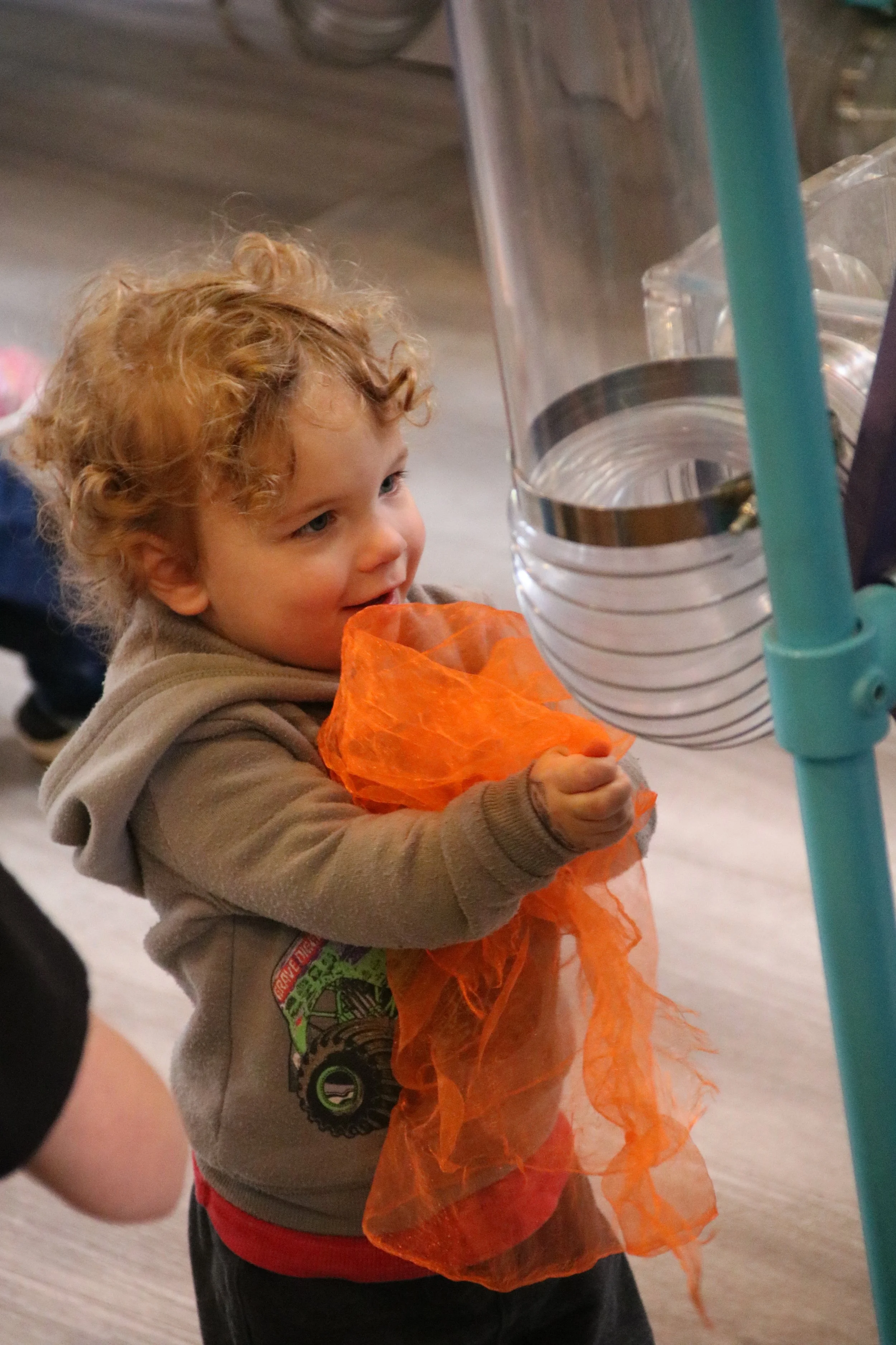 A young child with curly blonde hair wearing a beige hoodie and a red shirt, holding an orange mesh bag, looking at a water dispensing station.