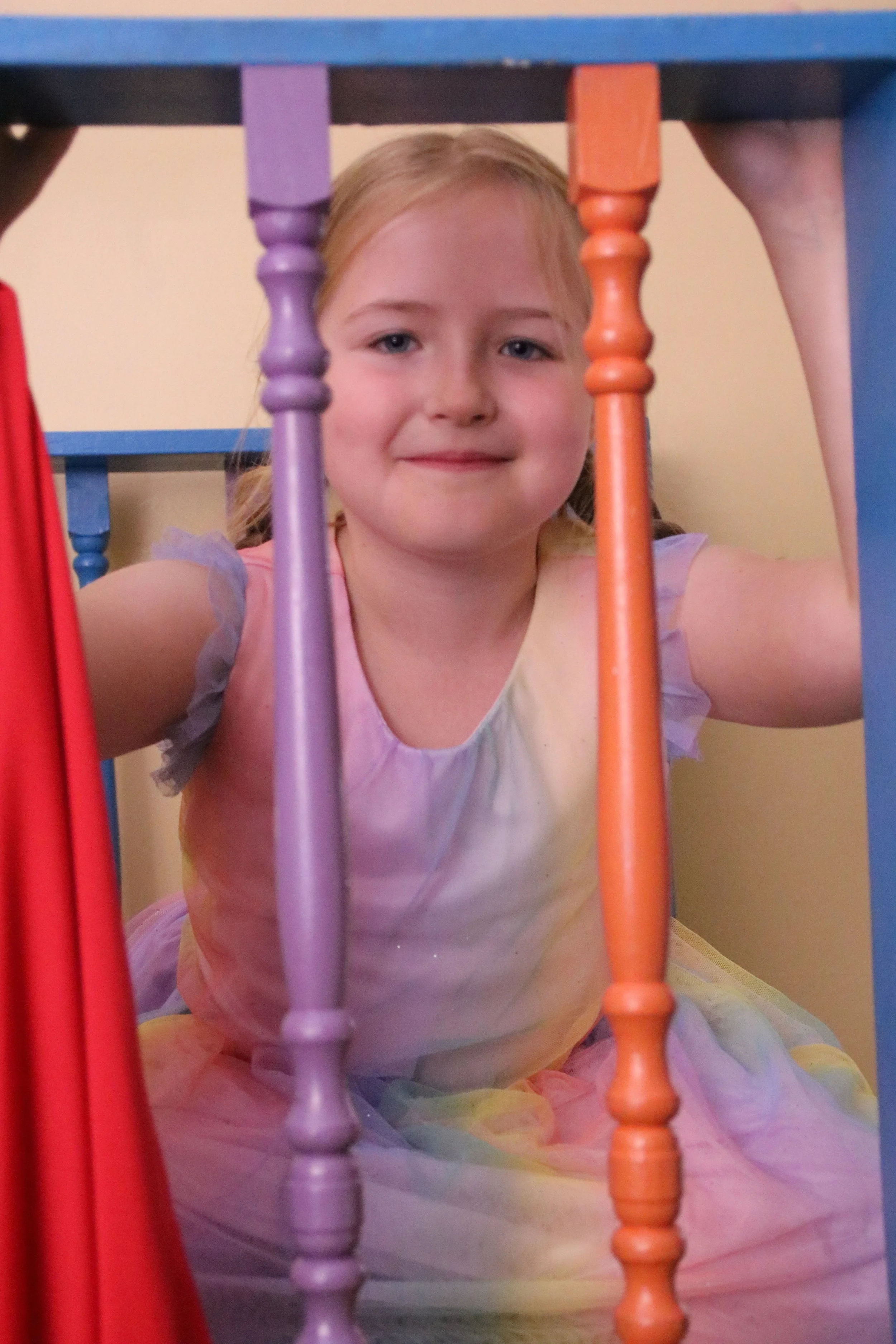 A young girl with blonde hair and a colorful dress looks through the wooden bars of a colorful play structure.