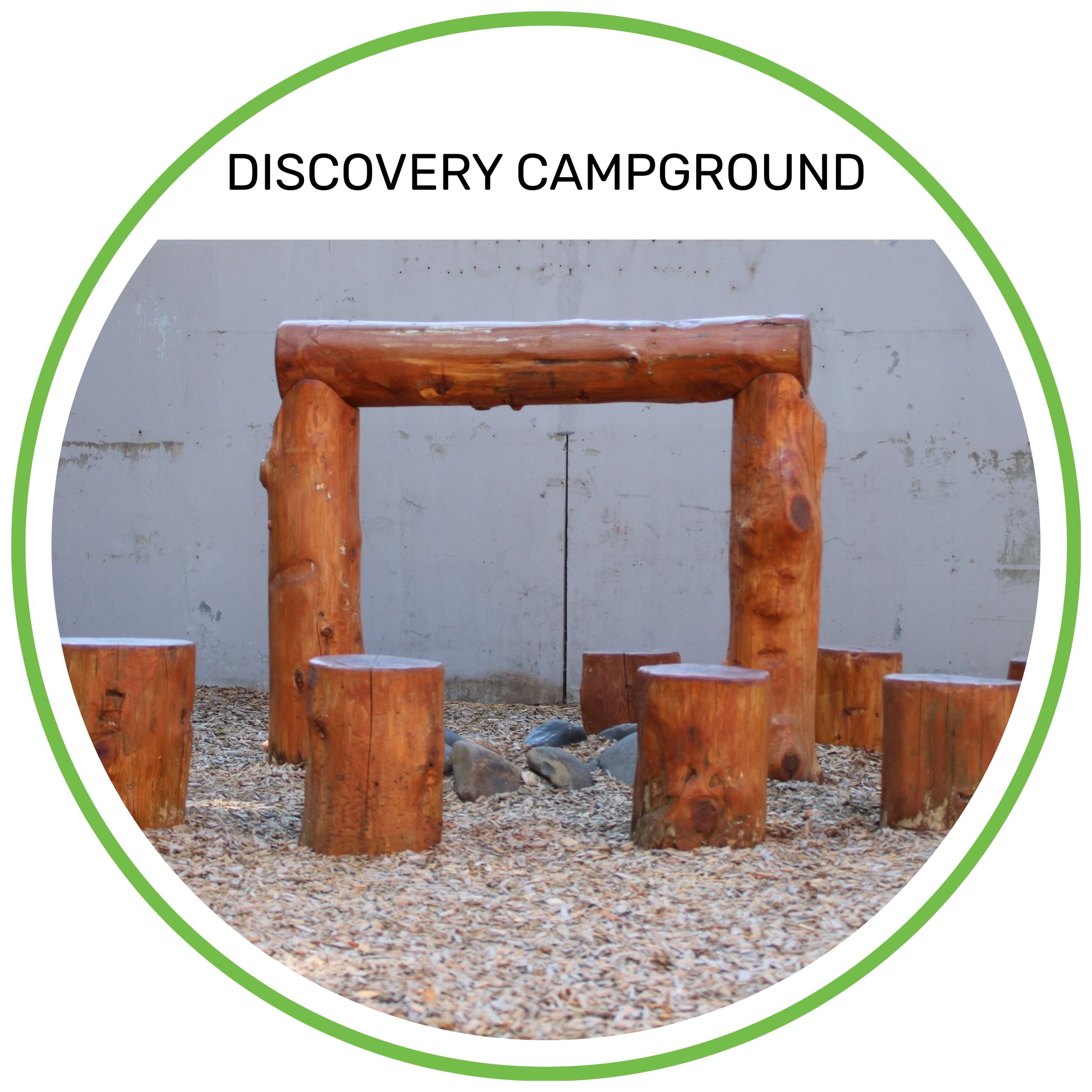 A wooden log playground structure labeled 'Discovery Campground', with logs arranged as benches and a frame, set on a gravel surface against a plain gray wall.