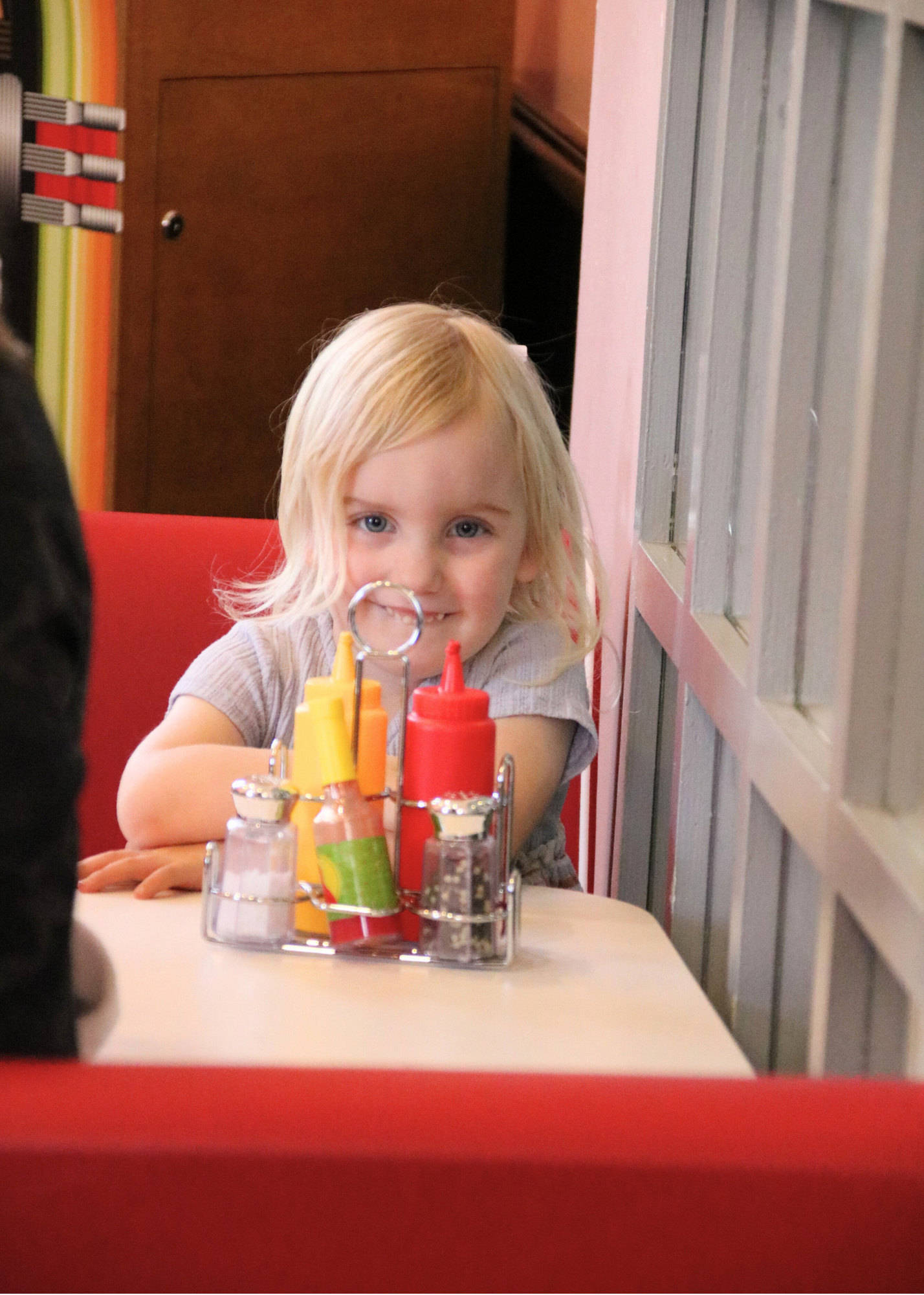 A young blonde girl sitting at a restaurant table, smiling and looking at the camera, with a condiment caddy containing ketchup, mustard, and salt and pepper shakers in front of her.